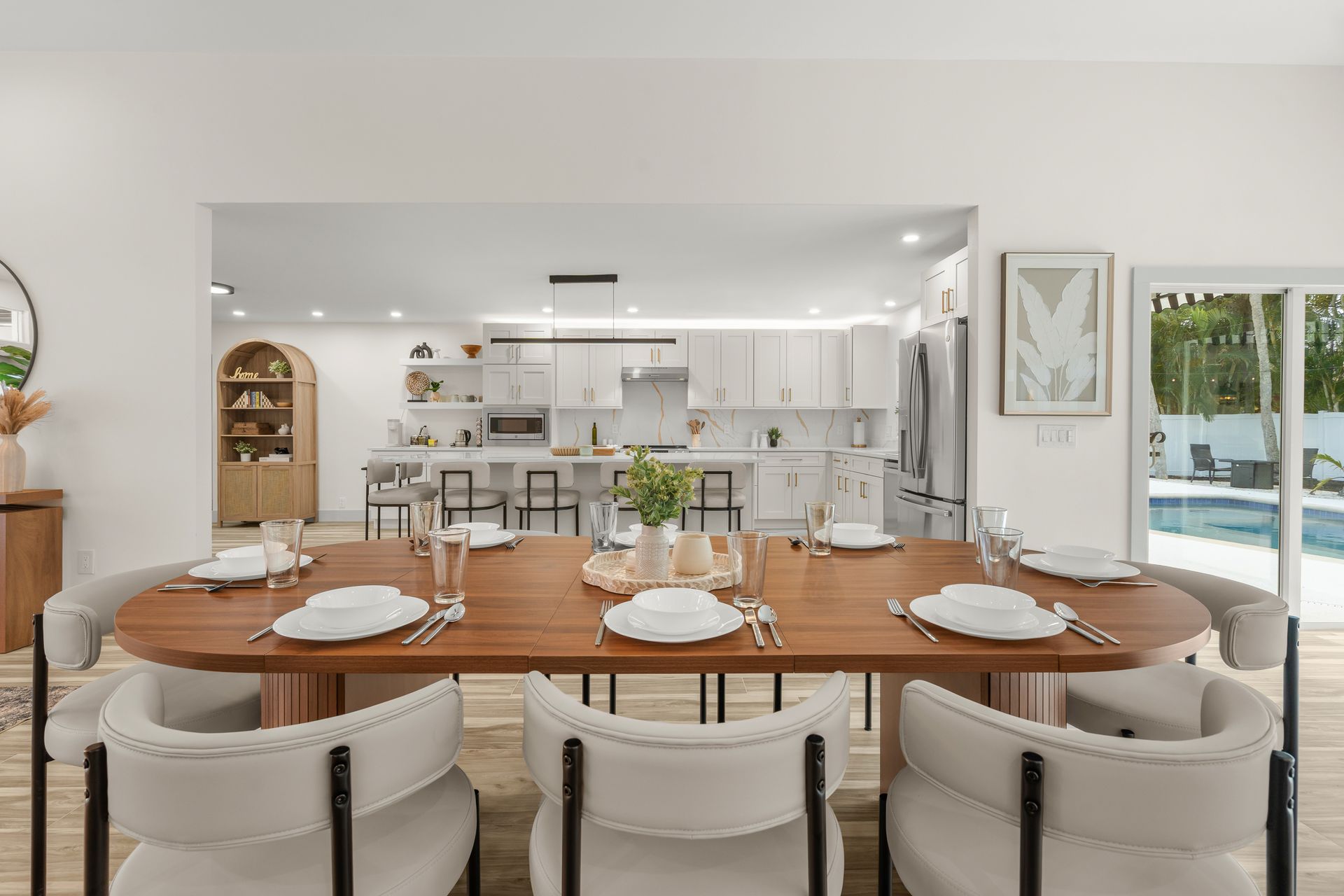 Modern dining room with a long wooden table set for a meal, opening to a bright kitchen and patio.
