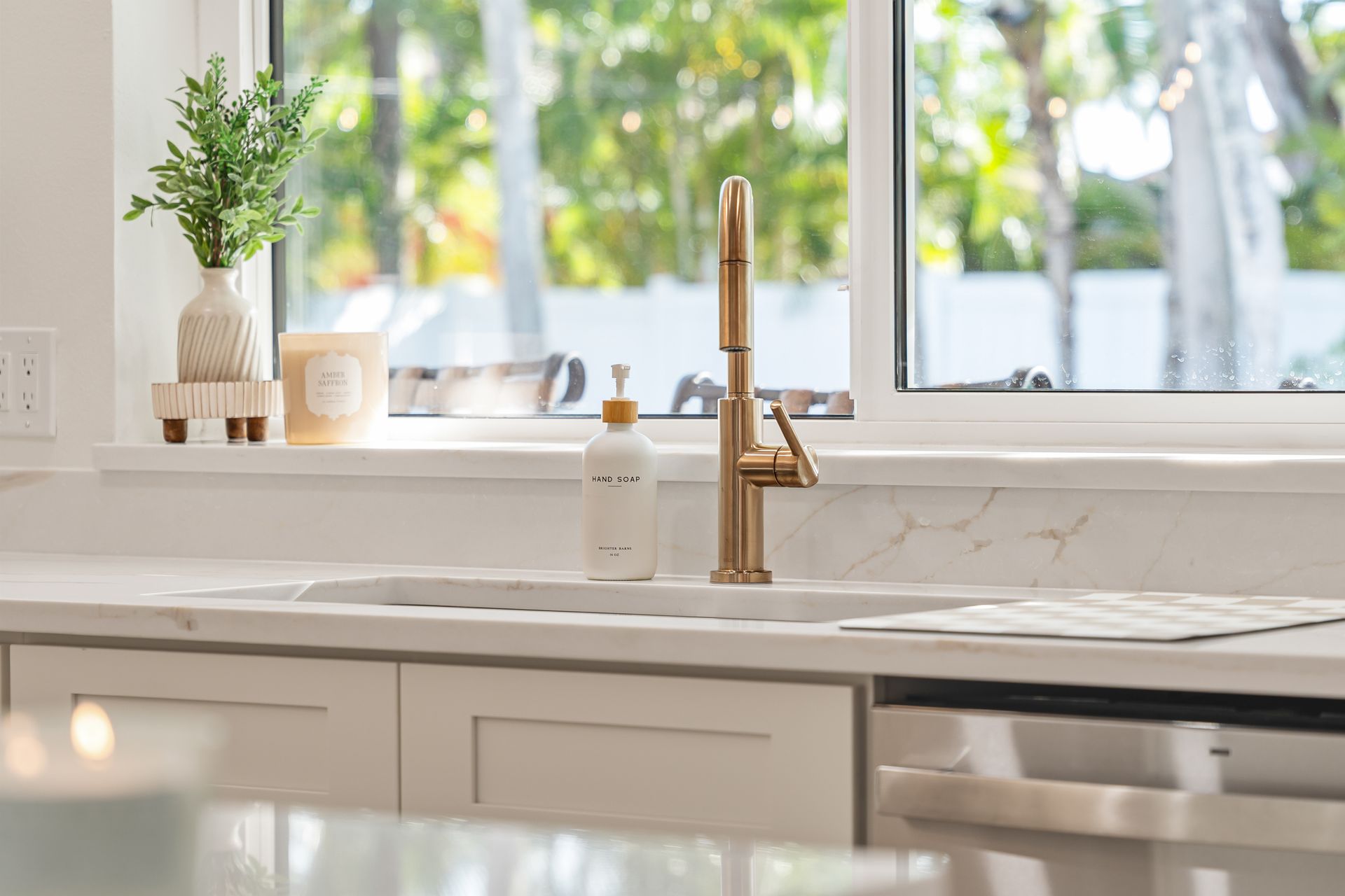 Bright kitchen sink with brass faucet, white marble countertop, and potted plant by a sunny window