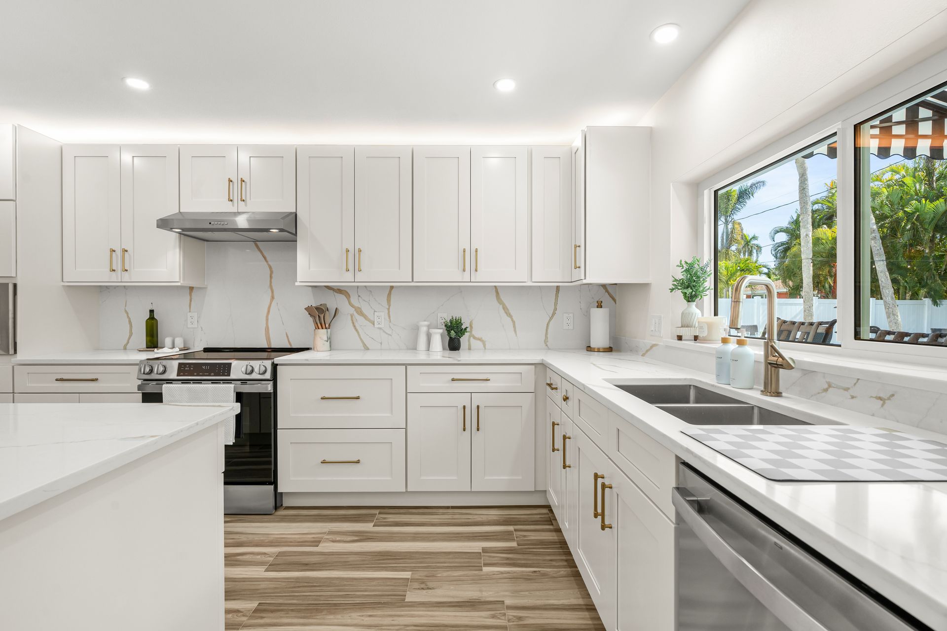 Bright modern white kitchen with stainless steel appliances, wood flooring, and a large window over the sink