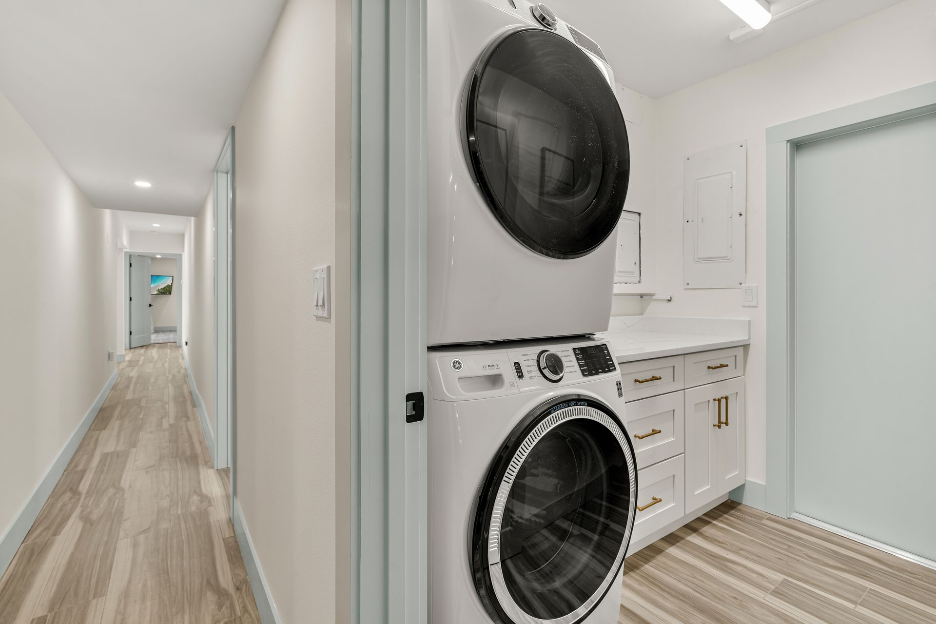 Bright hallway with stacked washer and dryer in a small laundry room, white cabinets, and light wood flooring