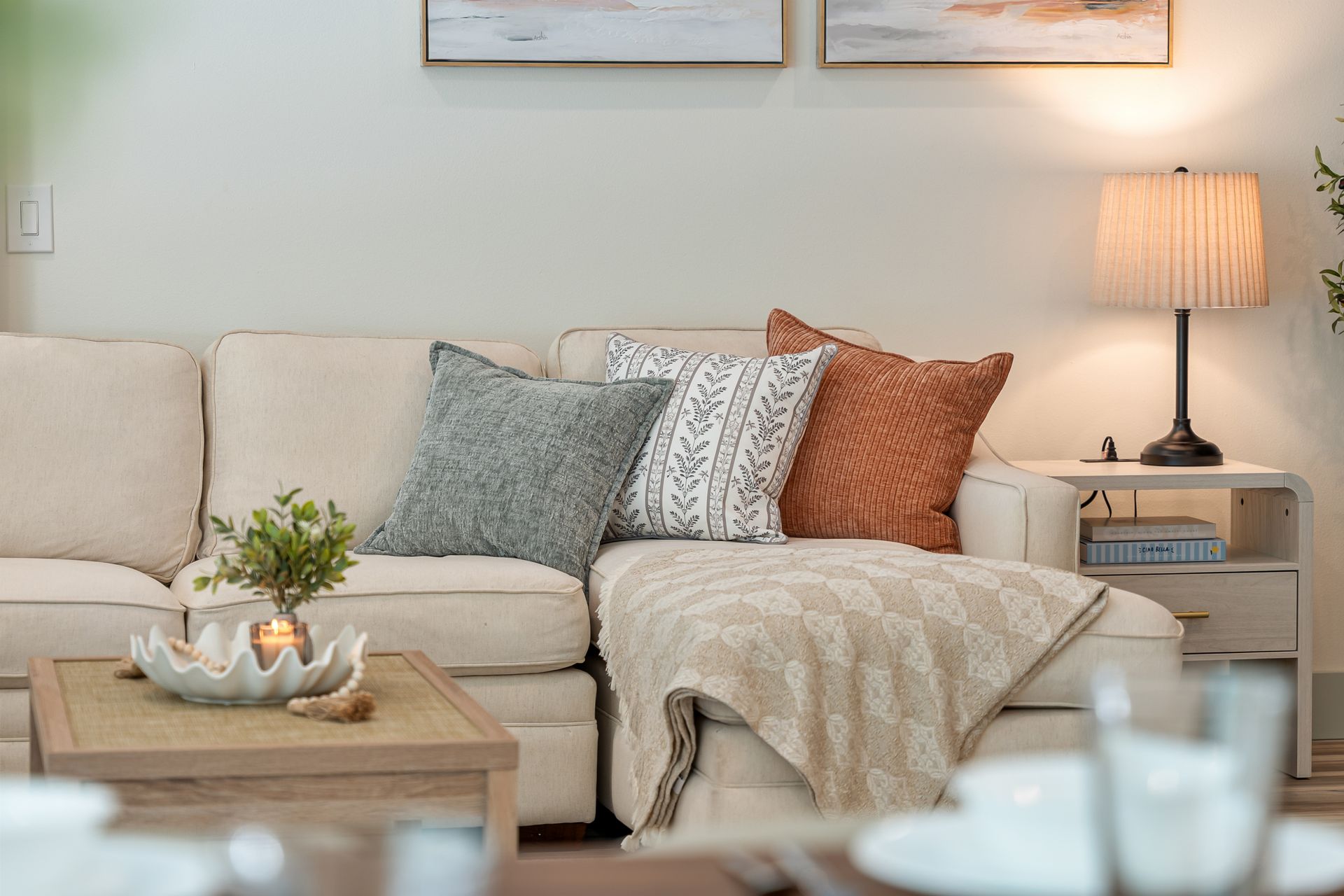 Cozy beige living room with sectional sofa, layered pillows, throw blanket, coffee table, and lit table lamp.