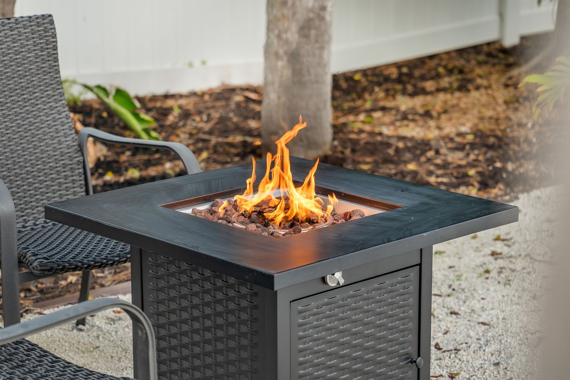Square black fire pit with flames on a patio beside outdoor chairs and a tree