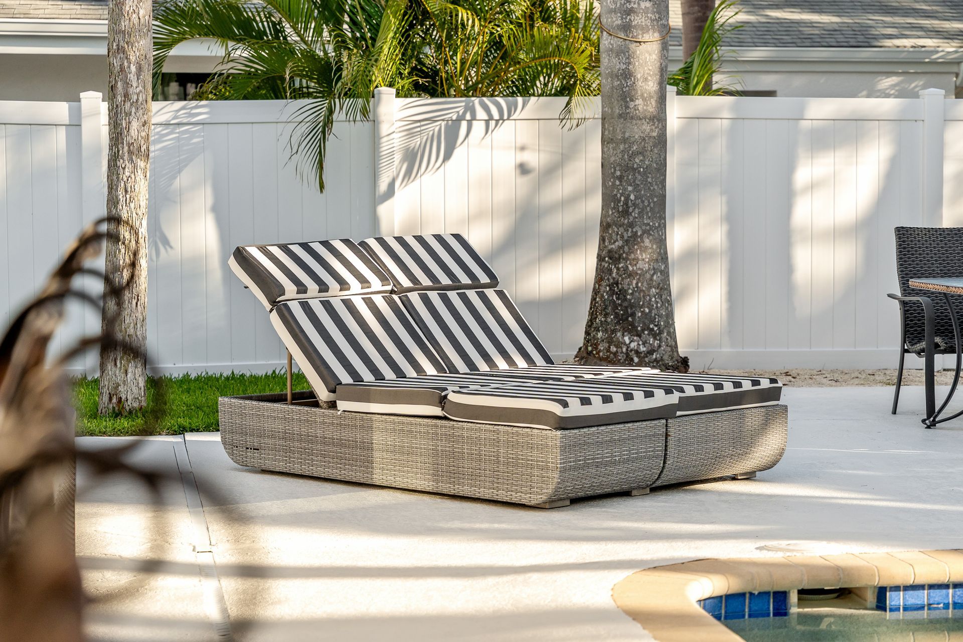 Striped chaise lounge by a pool in a sunny backyard with palm trees and a white fence