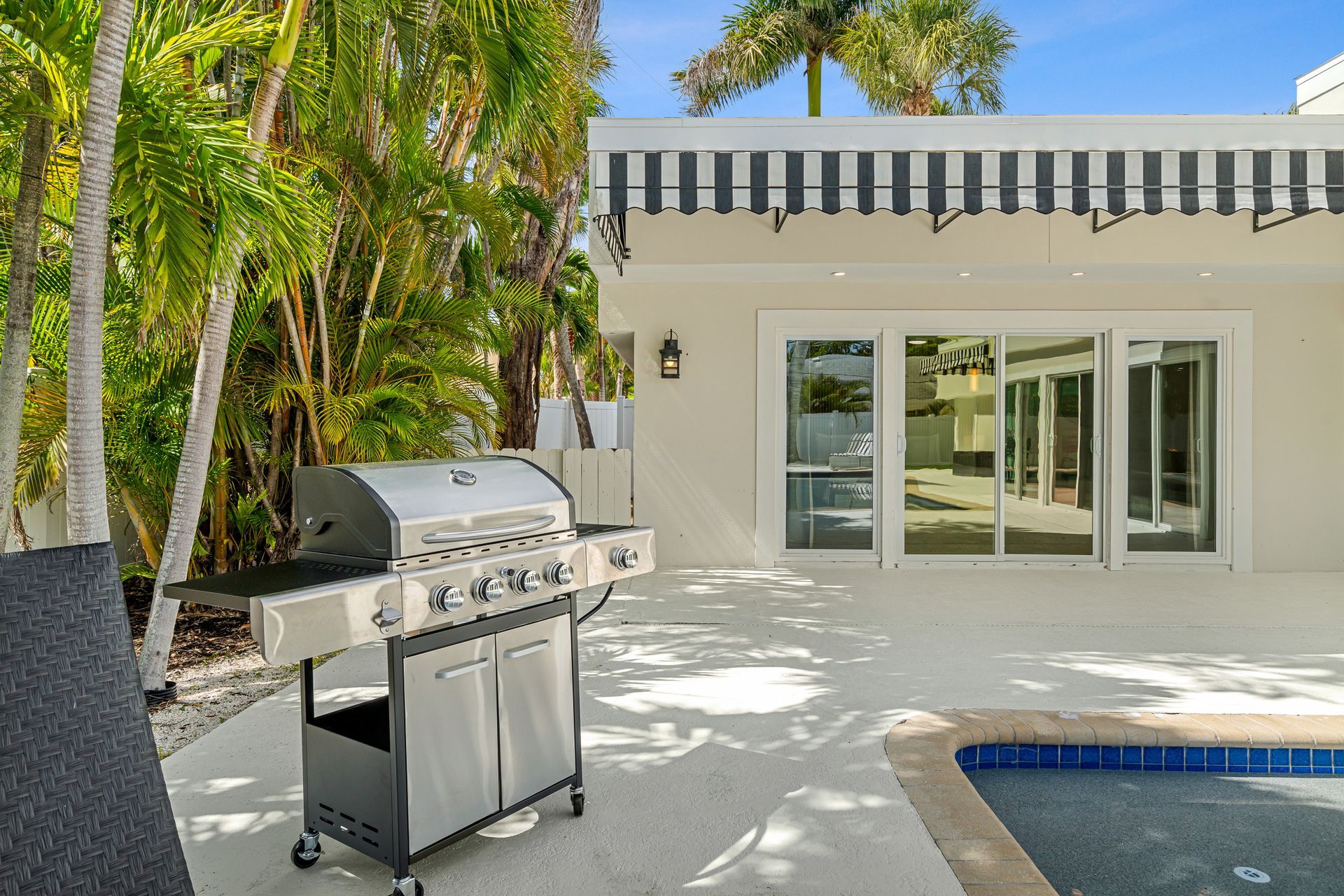 Patio with a stainless steel grill beside a pool and sliding glass doors, bordered by palm trees