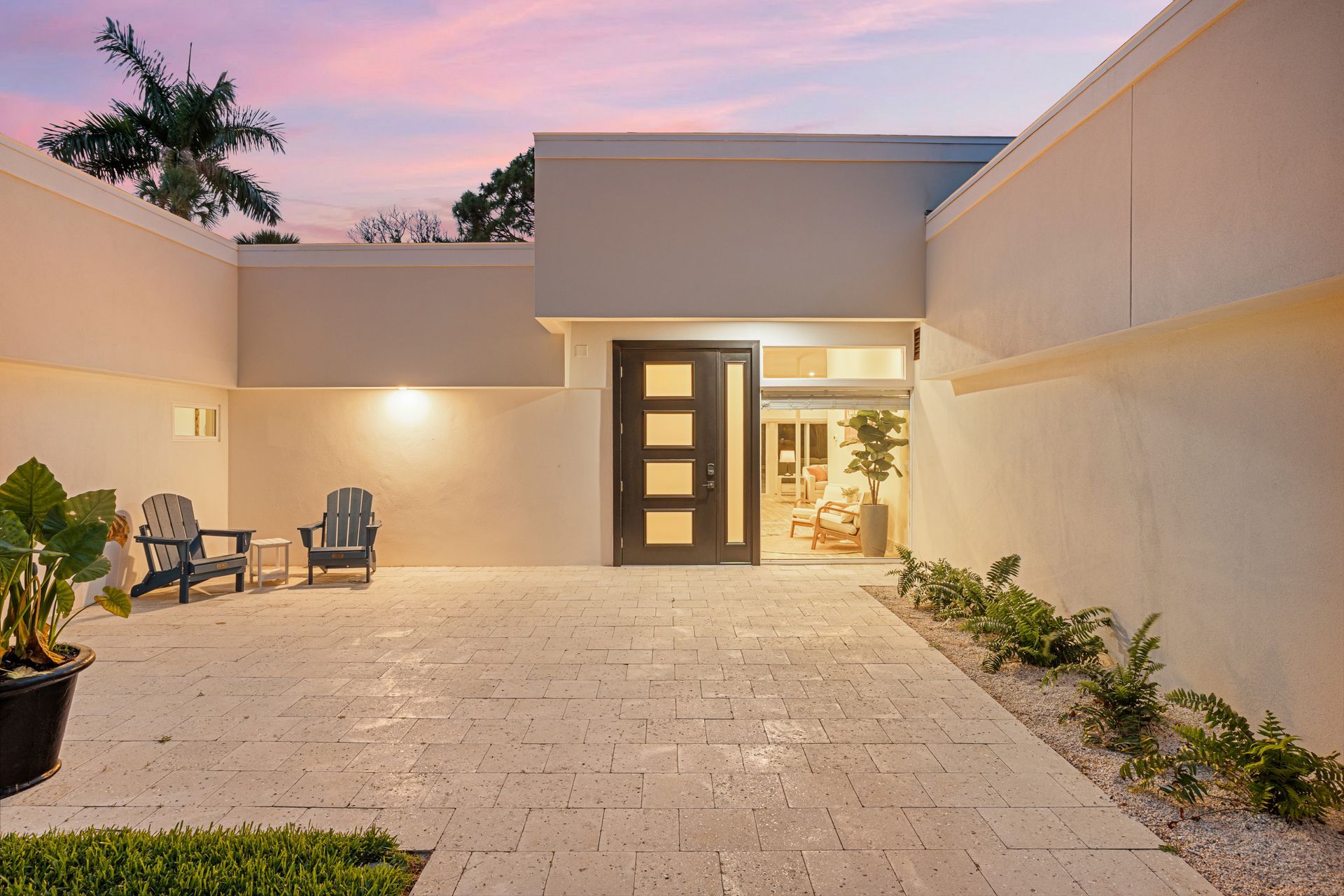 Modern courtyard patio at dusk with stone pavers, outdoor chairs, and an open front door lit from inside