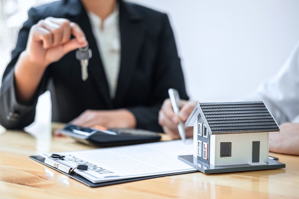 Persona con traje sosteniendo llaves sobre una casa en miniatura y papeles.