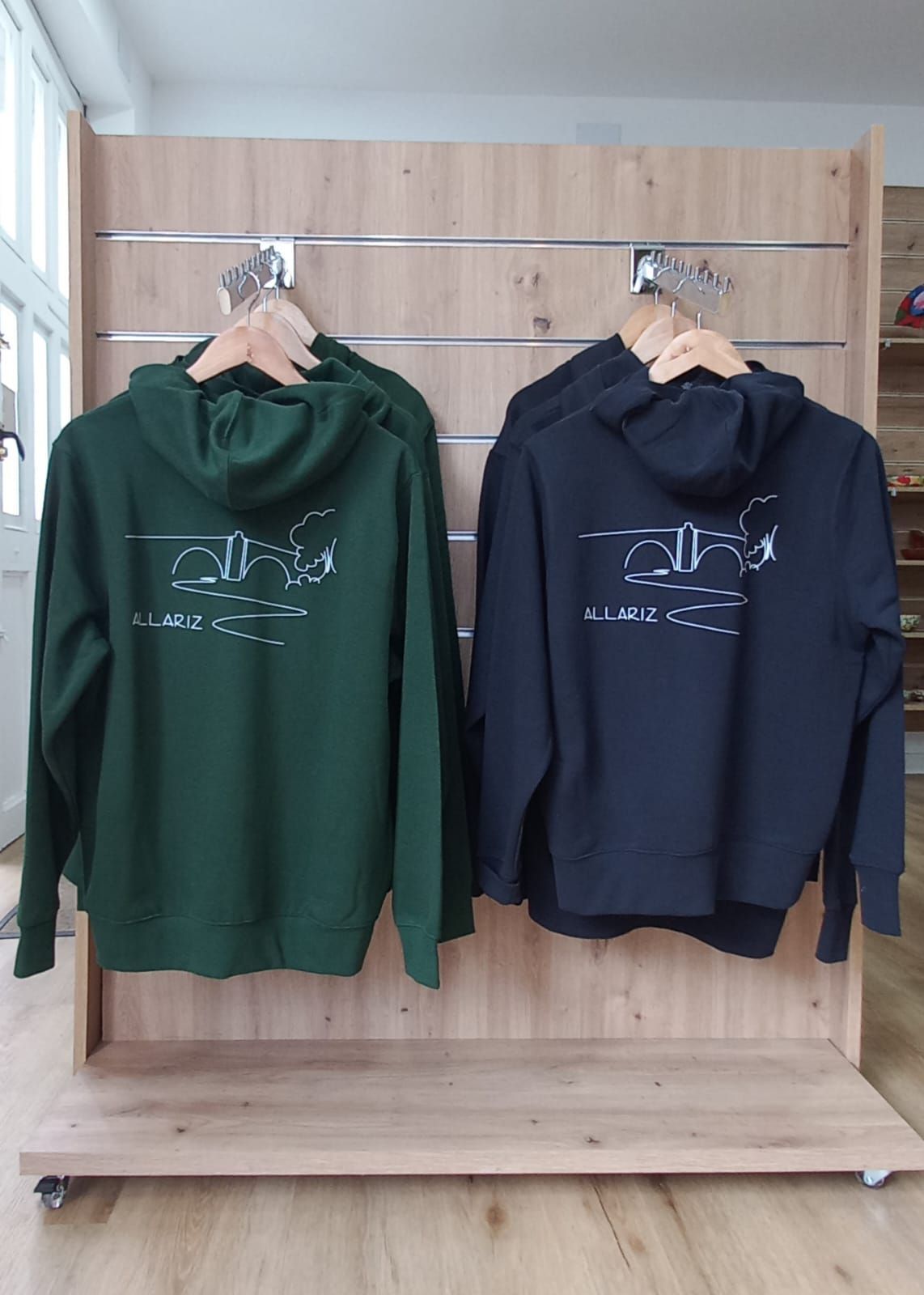 Dos sudaderas con capucha, una verde oscura y otra negra, colgadas en un expositor de madera. Las sudaderas tienen un logo blanco en la espalda.