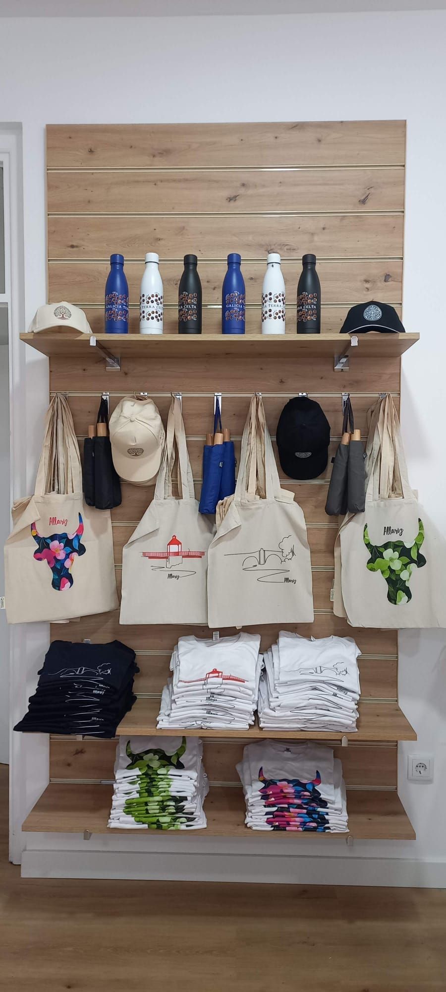 Expositor de madera con botellas, sombreros, bolsas de tela y camisetas dobladas.