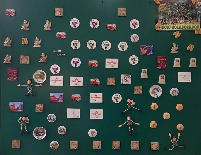 Tablón de anuncios verde cubierto con varios imanes, alfileres y pequeños elementos decorativos.