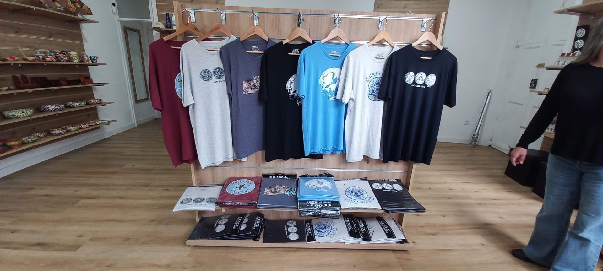 Exhibición de una tienda de ropa con camisetas de varios colores y diseños. Se ve una persona a la derecha.