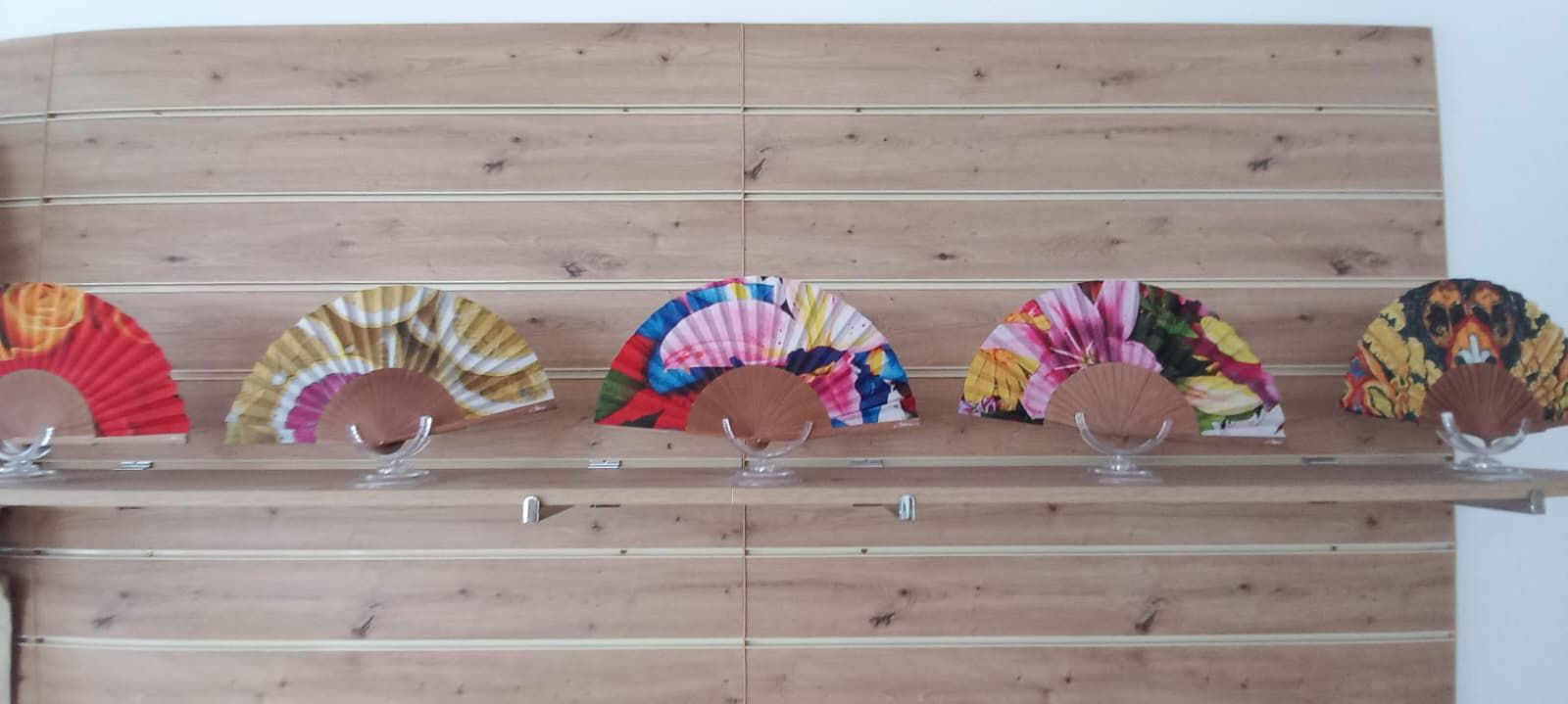 Cinco abanicos decorativos exhibidos en un estante de madera, cada uno con un patrón colorido diferente.