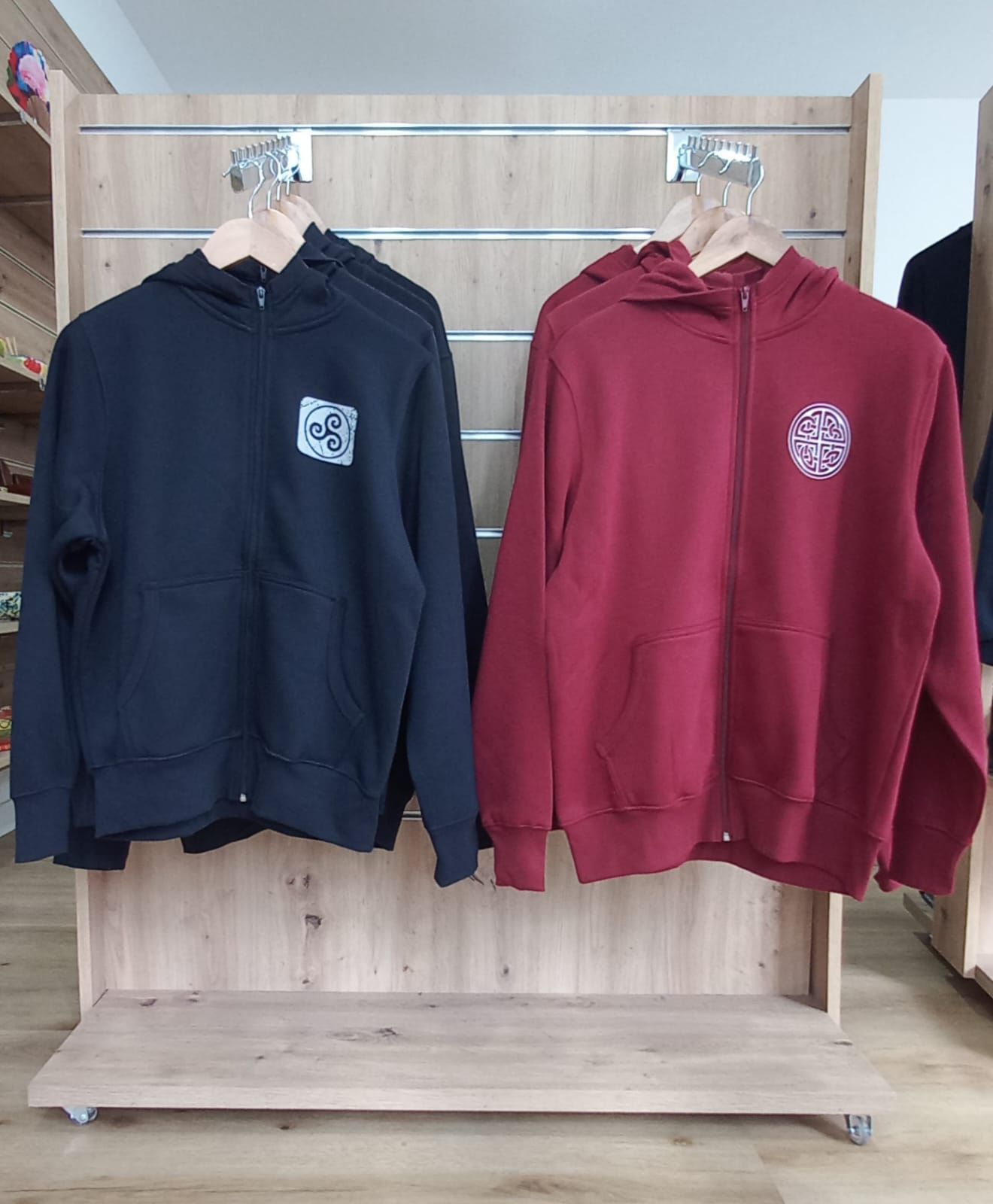 Sudadera con capucha, negra y roja, con cremallera y logotipos colgados en un expositor de madera.