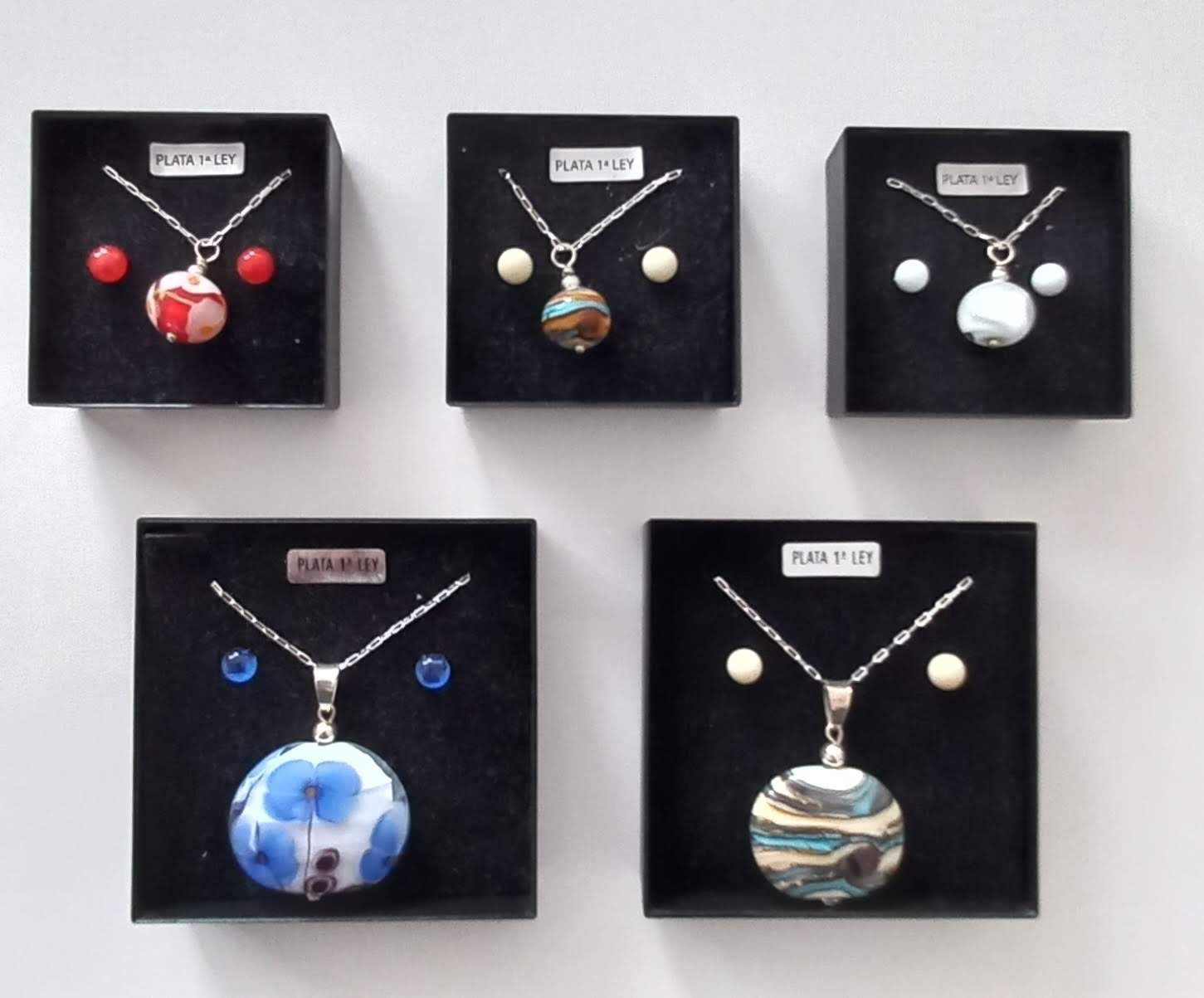 Cinco collares con aretes a juego en cajas negras, que muestran diseños de colgantes de vidrio coloridos.