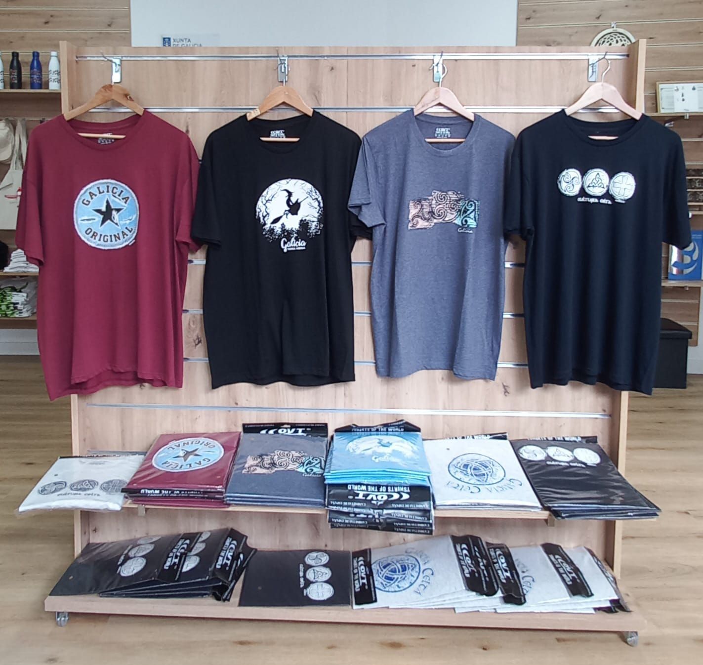 Cuatro camisetas colgadas en un expositor de madera con camisetas dobladas debajo.