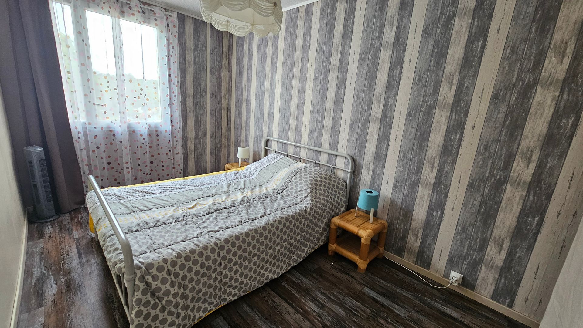 Chambre avec lit, table de chevet, papier peint à motifs et parquet. Fenêtre avec rideaux.