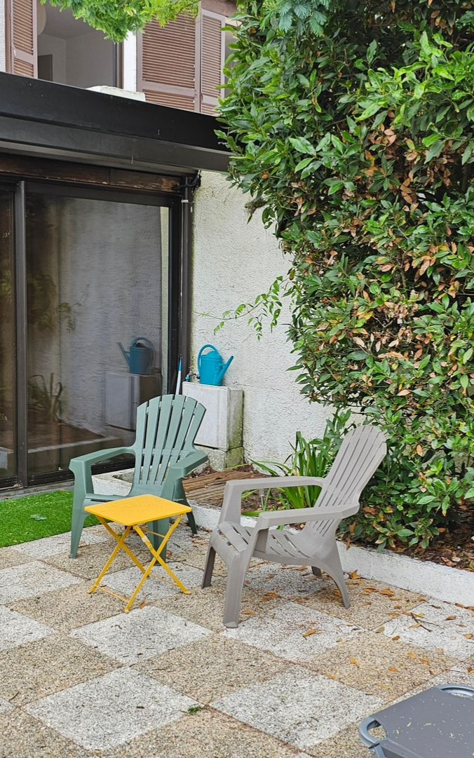 Terrasse avec deux chaises en plastique, une petite table et un grand buisson. Deux arrosoirs sont visibles.