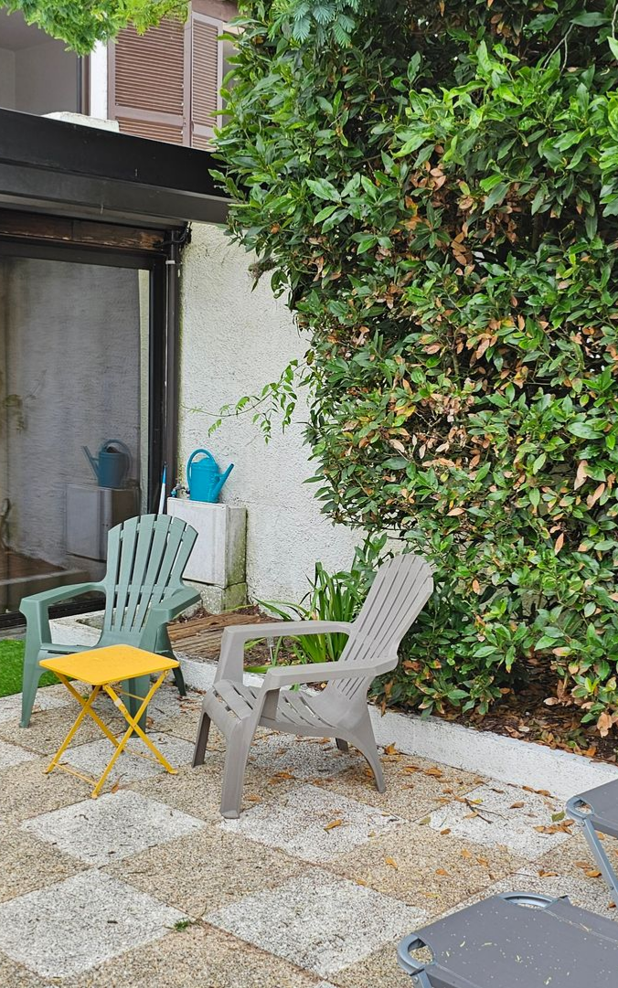 Terrasse avec deux chaises en plastique, une petite table jaune et une végétation luxuriante.
