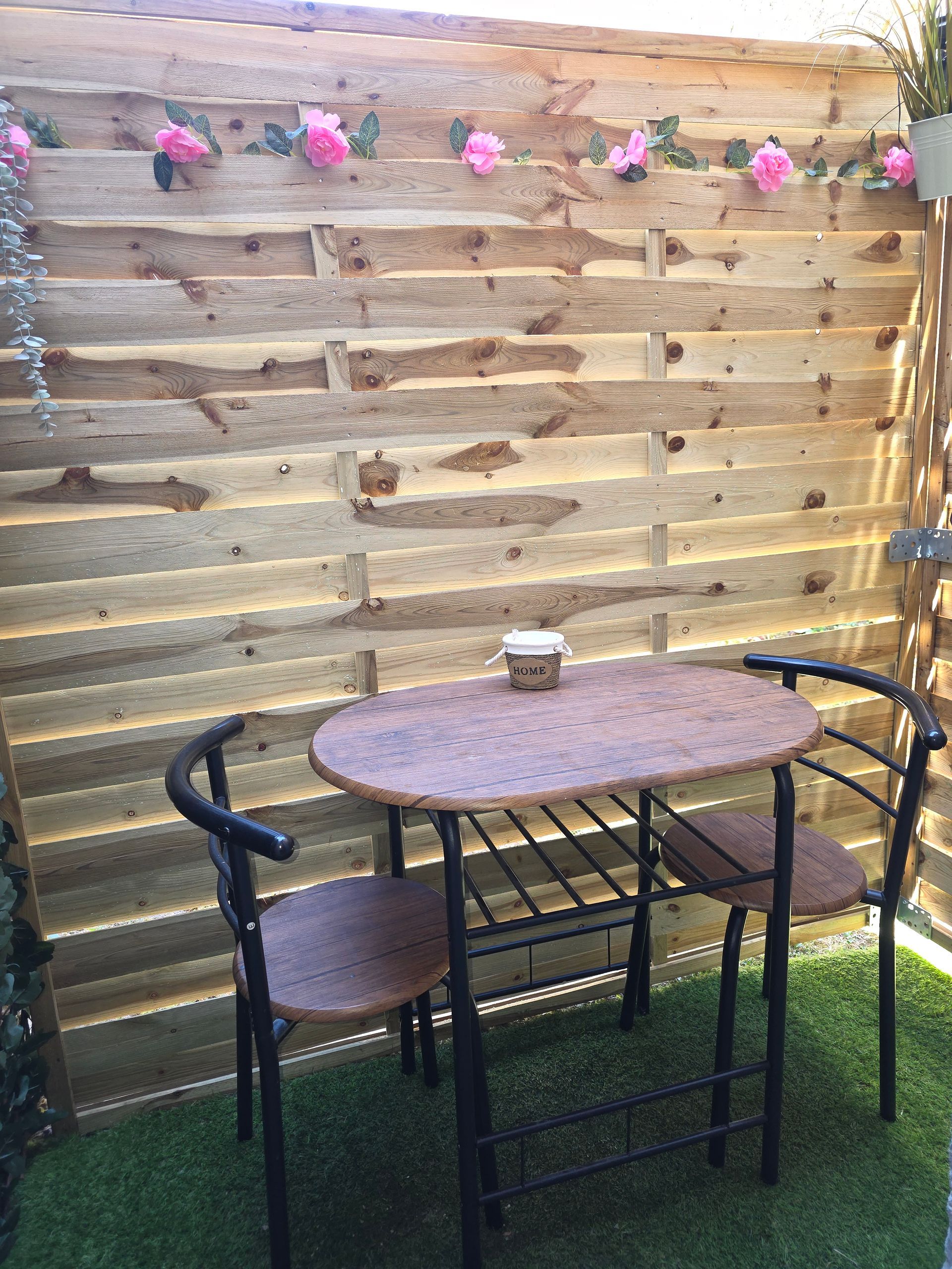 Terrasse avec deux chaises en plastique, une petite table et un grand buisson. Deux arrosoirs sont visibles.