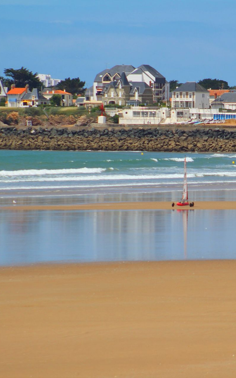 Scène de plage avec un char à voile sur le sable, une ville en arrière-plan, des vagues et un ciel bleu.