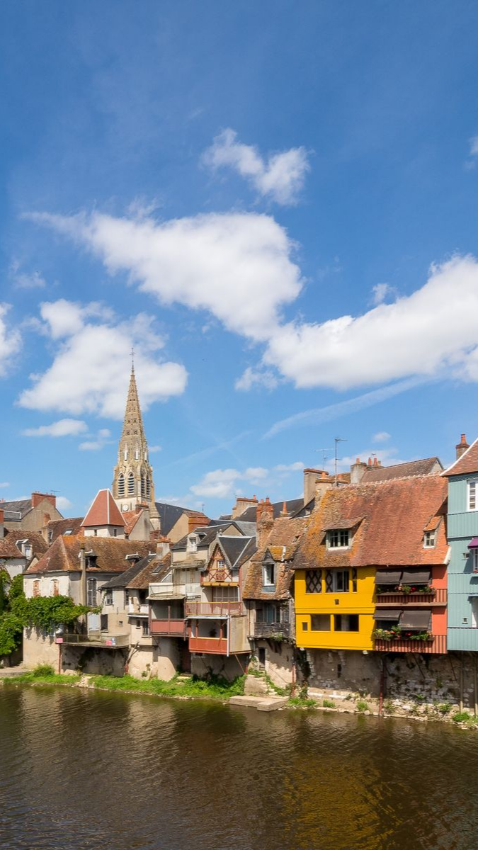 Des bâtiments colorés bordent une rivière sous un ciel bleu, avec le clocher d'une haute église en arrière-plan.