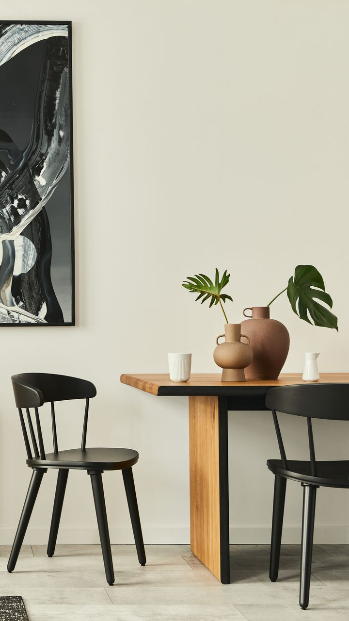 Salle à manger avec table en bois, chaises noires et vases à feuilles vertes. Œuvre d'art en noir et blanc au mur.