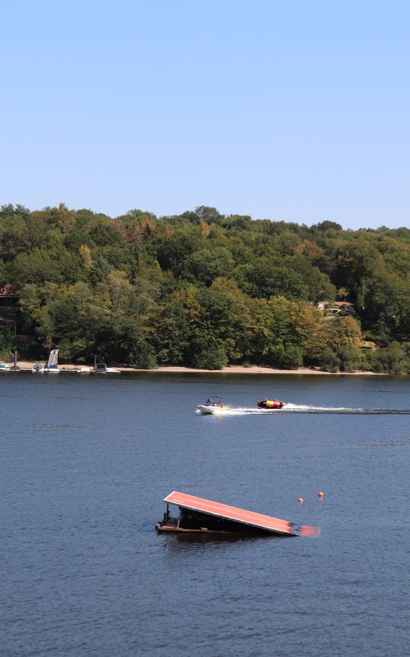Scène de lac avec un bateau tirant une bouée, un quai, des arbres en arrière-plan, un ciel bleu.