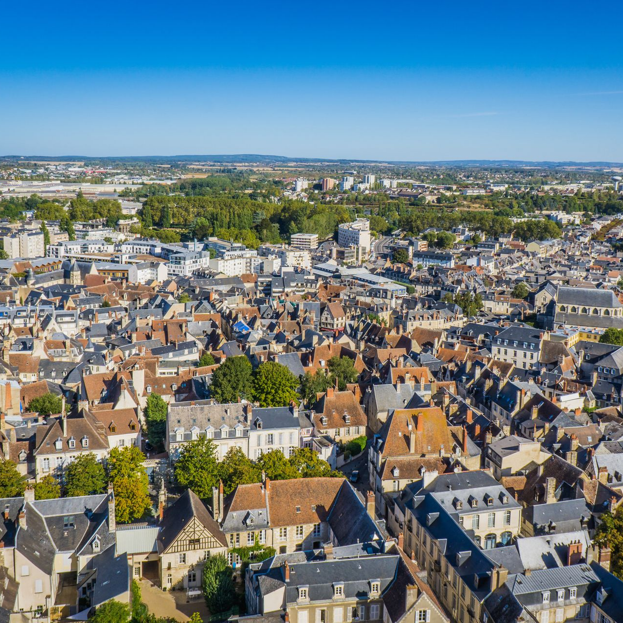 Vue aérienne d'une ville aux toits de tuiles rouges, aux bâtiments imposants et à la végétation luxuriante se détachant sur un ciel bleu.