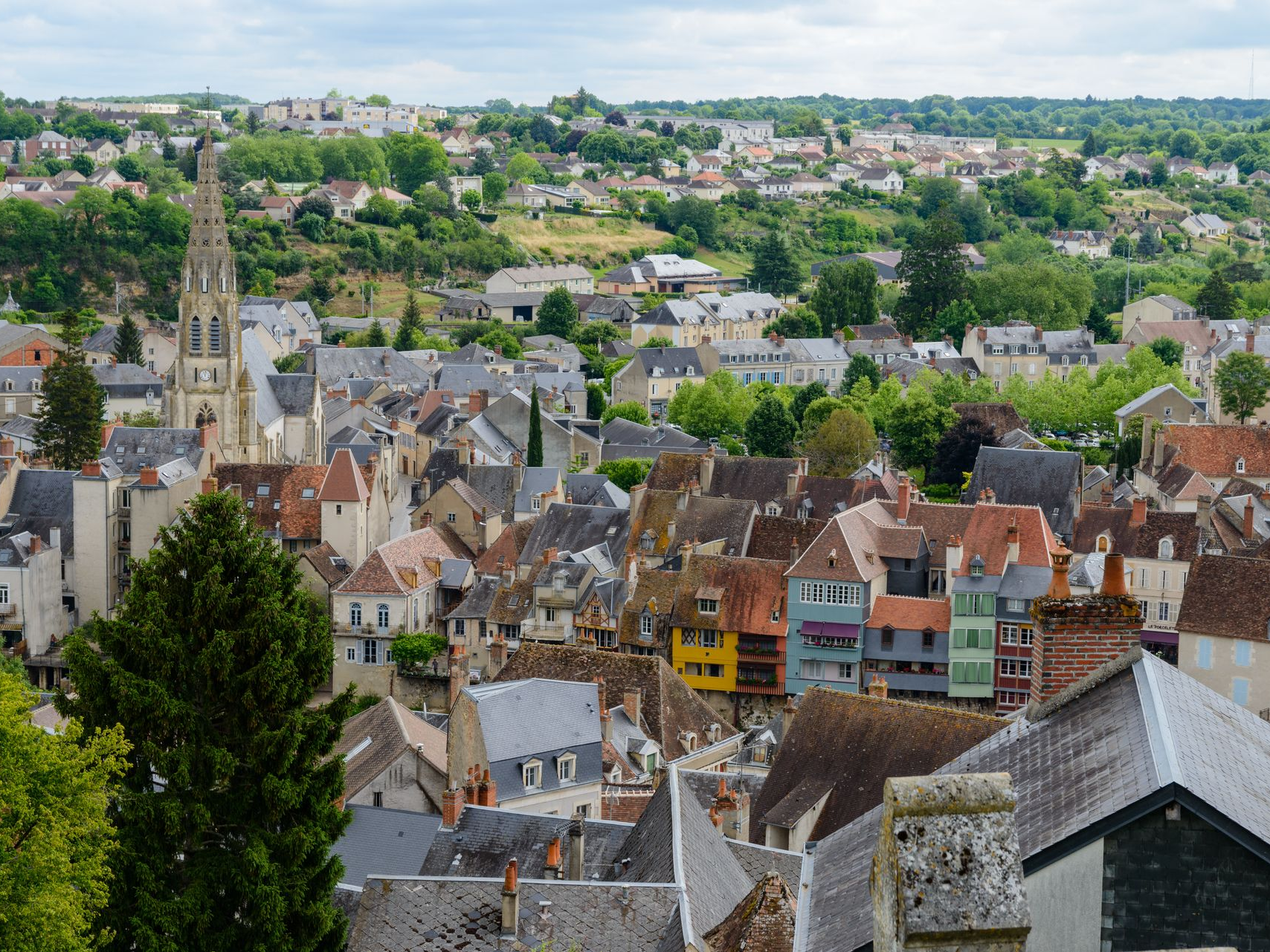 Vue d'une ville européenne avec des bâtiments colorés, des toits et un clocher, le tout se détachant sur un fond d'arbres verdoyants sous un ciel nuageux.