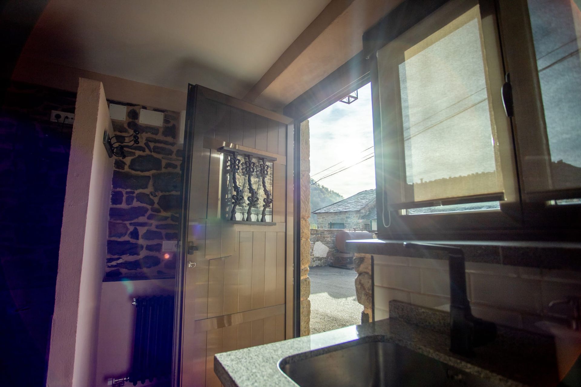 Puerta interior con luminosa vista exterior, pared de piedra, lavabo y ventana.