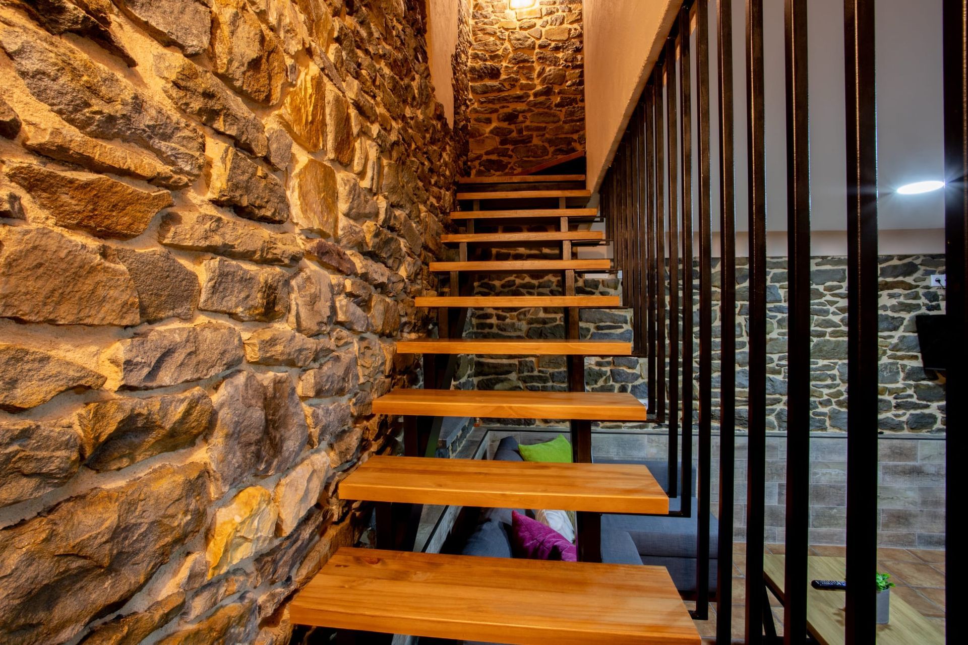Escalera de pared de piedra con escalones de madera y barandilla de metal negro; iluminación cálida.