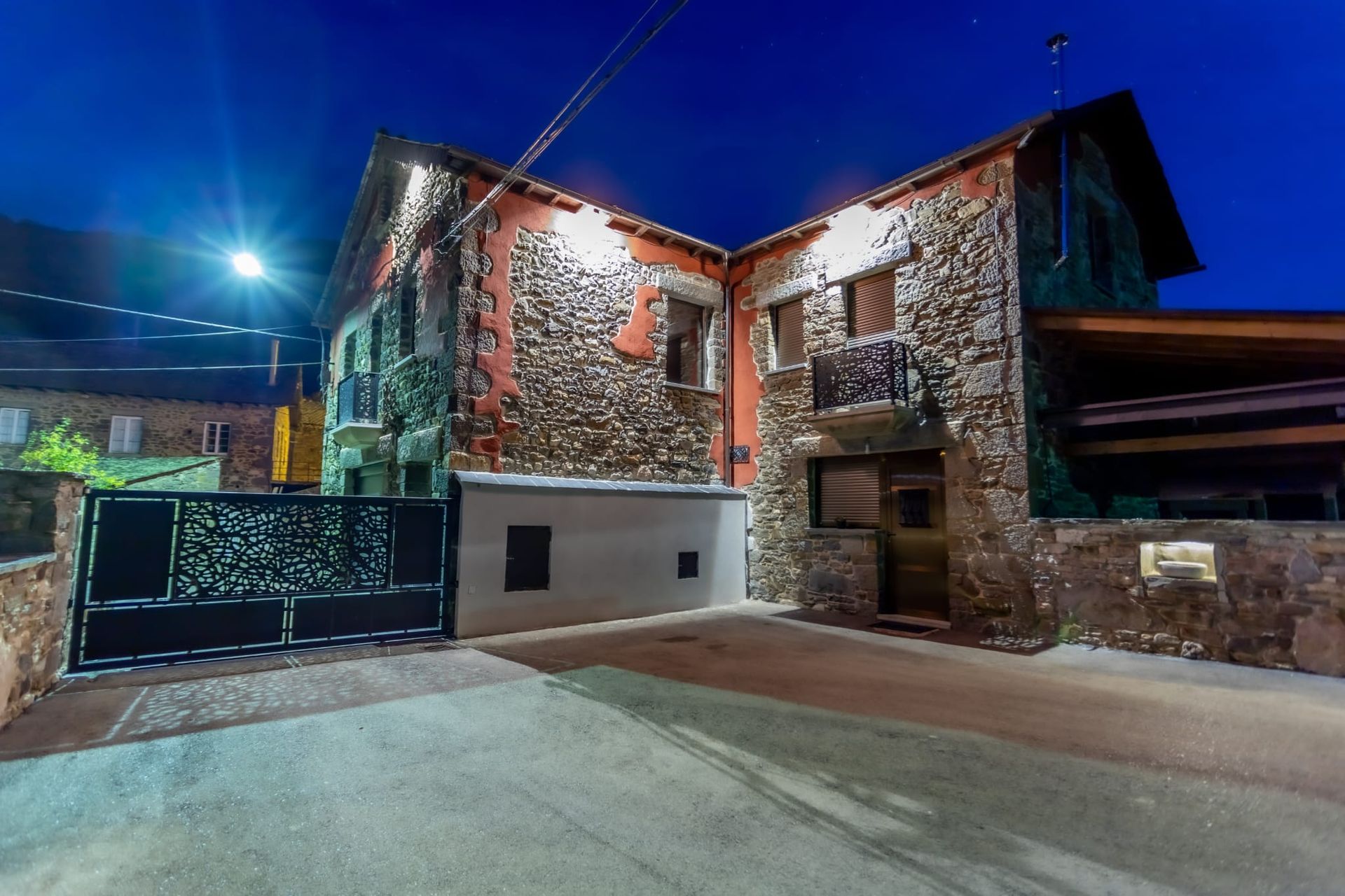 Casa de piedra al anochecer, iluminada por farolas. Puerta de hierro forjado negra, cielo azul.