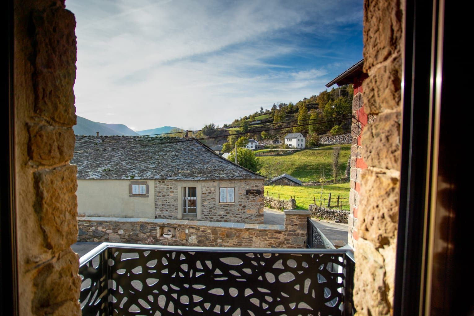 Vista a través de una ventana de piedra: casas rurales, colinas verdes y un balcón de metal estampado.