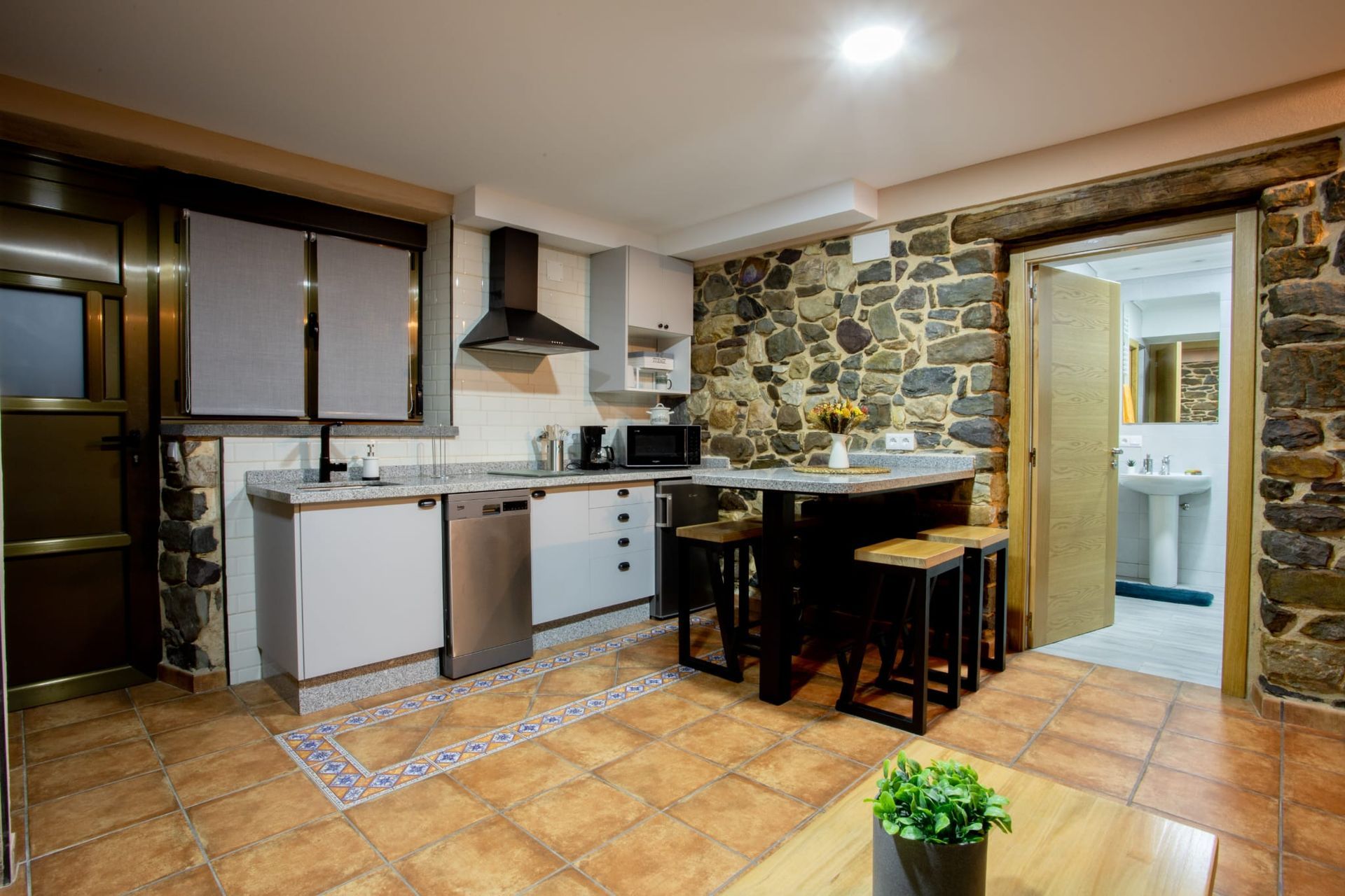 Cocina con paredes de piedra, gabinetes blancos, electrodomésticos de acero inoxidable, barra desayunadora y puerta abierta.