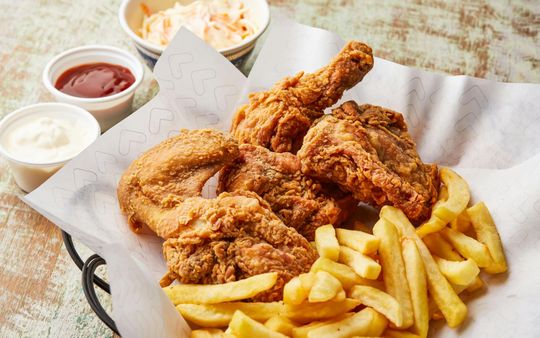 Trozos de pollo frito crujiente y patatas fritas en una cesta con ensalada de col y salsas para mojar sobre una superficie rústica de madera.