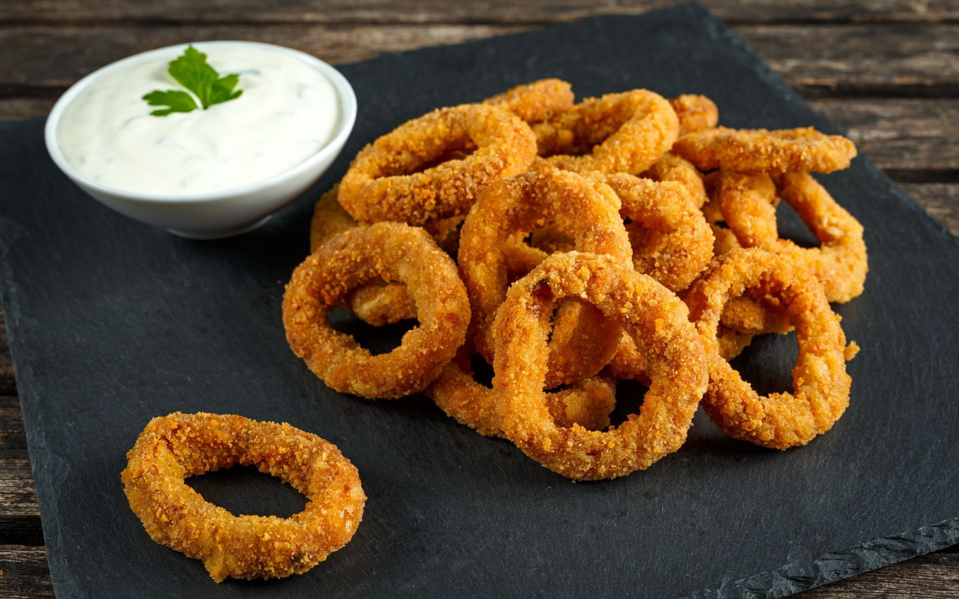 Aros de cebolla fritos y dorados apilados sobre una pizarra oscura, con un bol de salsa blanca para mojar adornado con perejil.