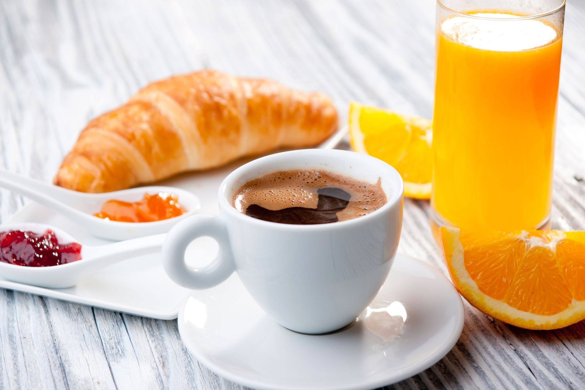 Desayuno: croissant, café, zumo de naranja, mermeladas y rodajas de naranja sobre una mesa de madera blanca.