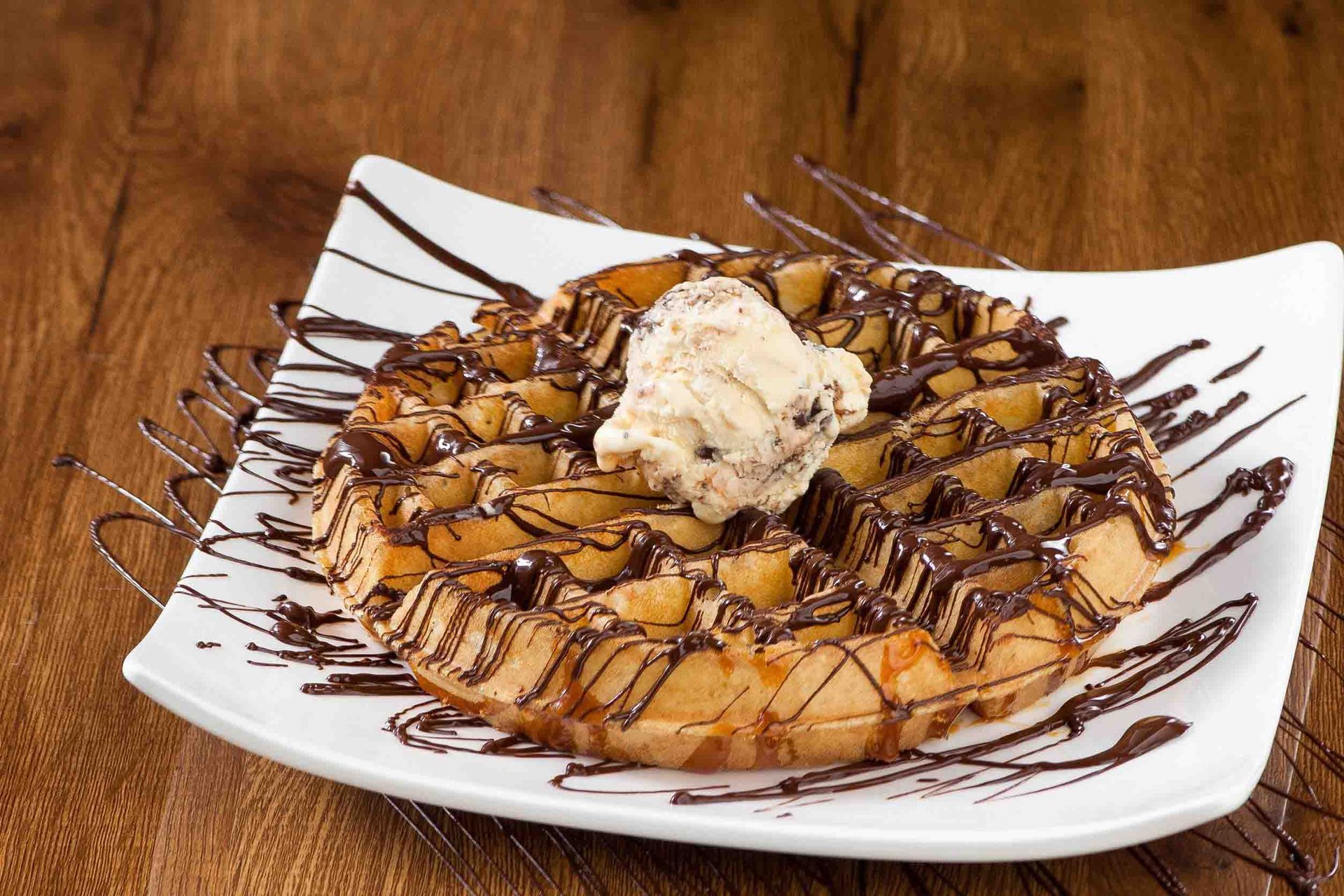 Gofre cubierto con salsa de chocolate y helado, servido en un plato blanco sobre un fondo de madera.
