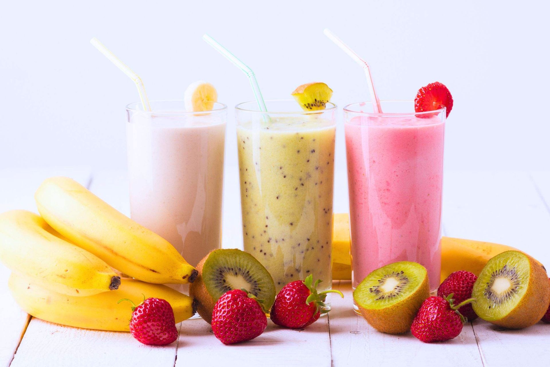 Tres batidos de frutas con pajitas, junto con plátanos frescos, kiwis y fresas.