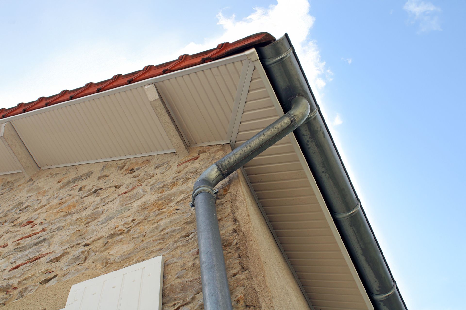 Vue plongeante sur le toit d'une maison avec gouttière, descente pluviale et bardage beige sur fond de ciel bleu.