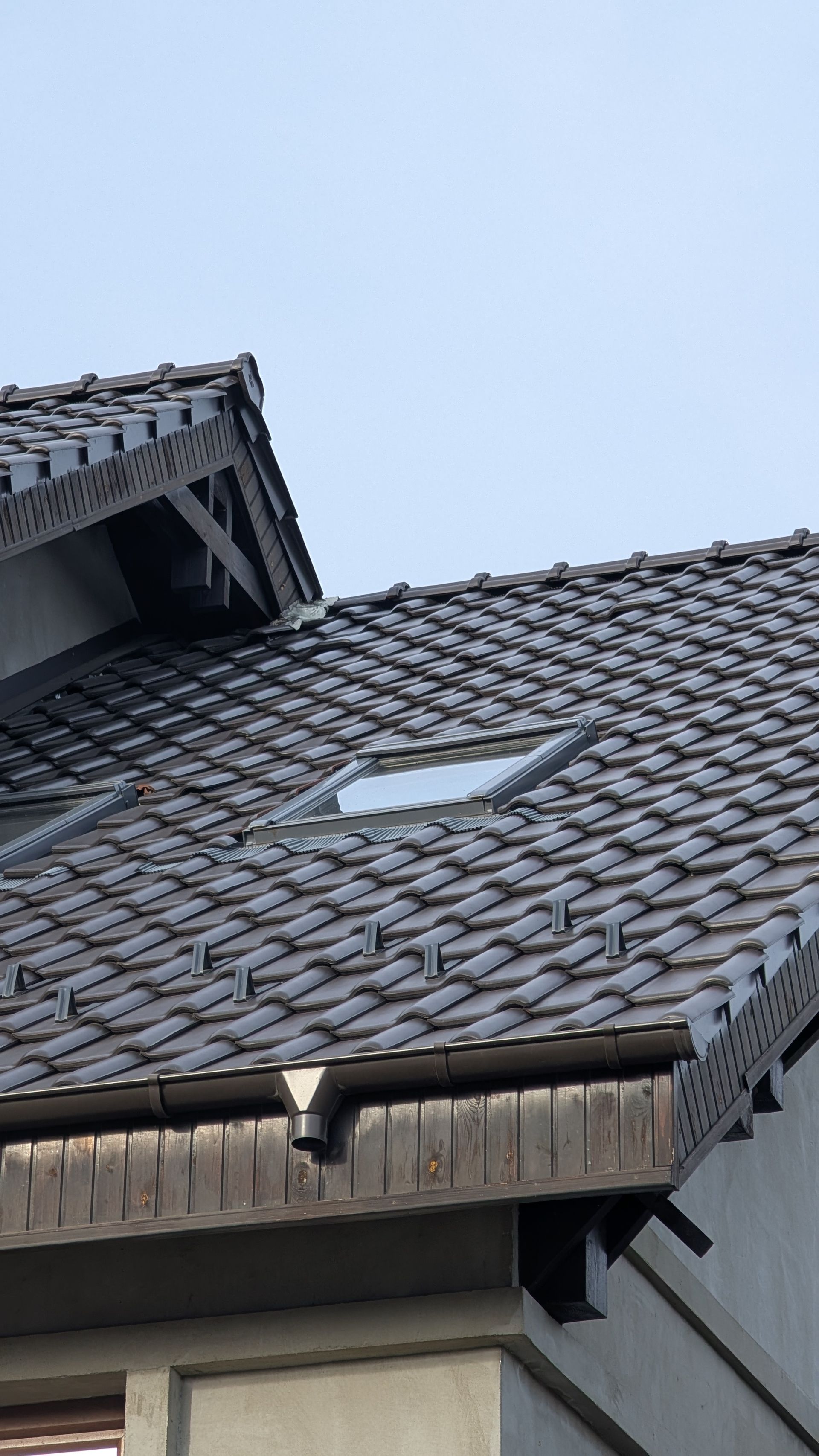 Toiture en tuiles grises avec une fenêtre de toit intégrée, un pignon en bois et une gouttière sous un ciel gris.