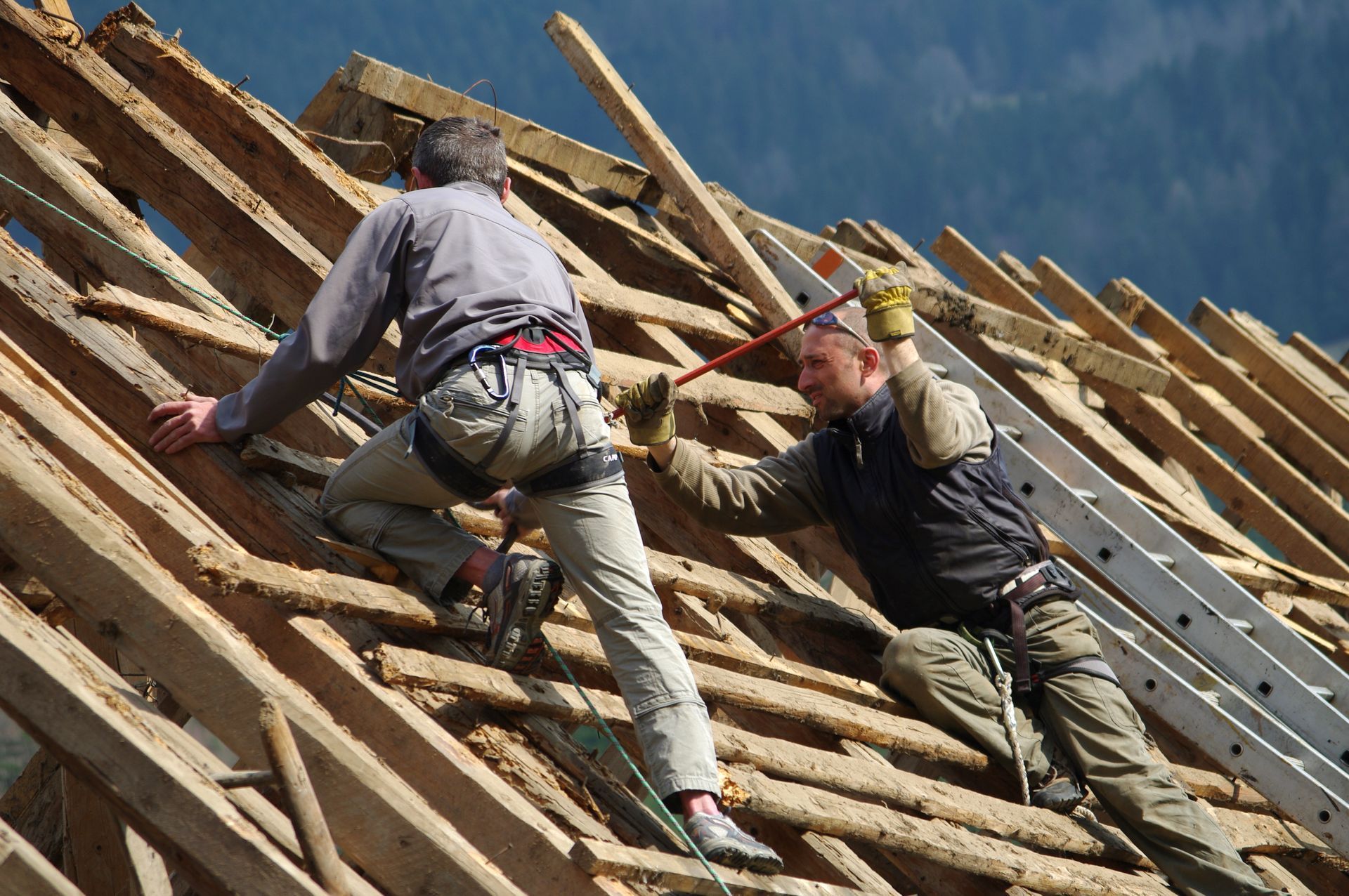 Deux ouvriers escaladent une charpente de toit en bois abrupte sous un ciel bleu.