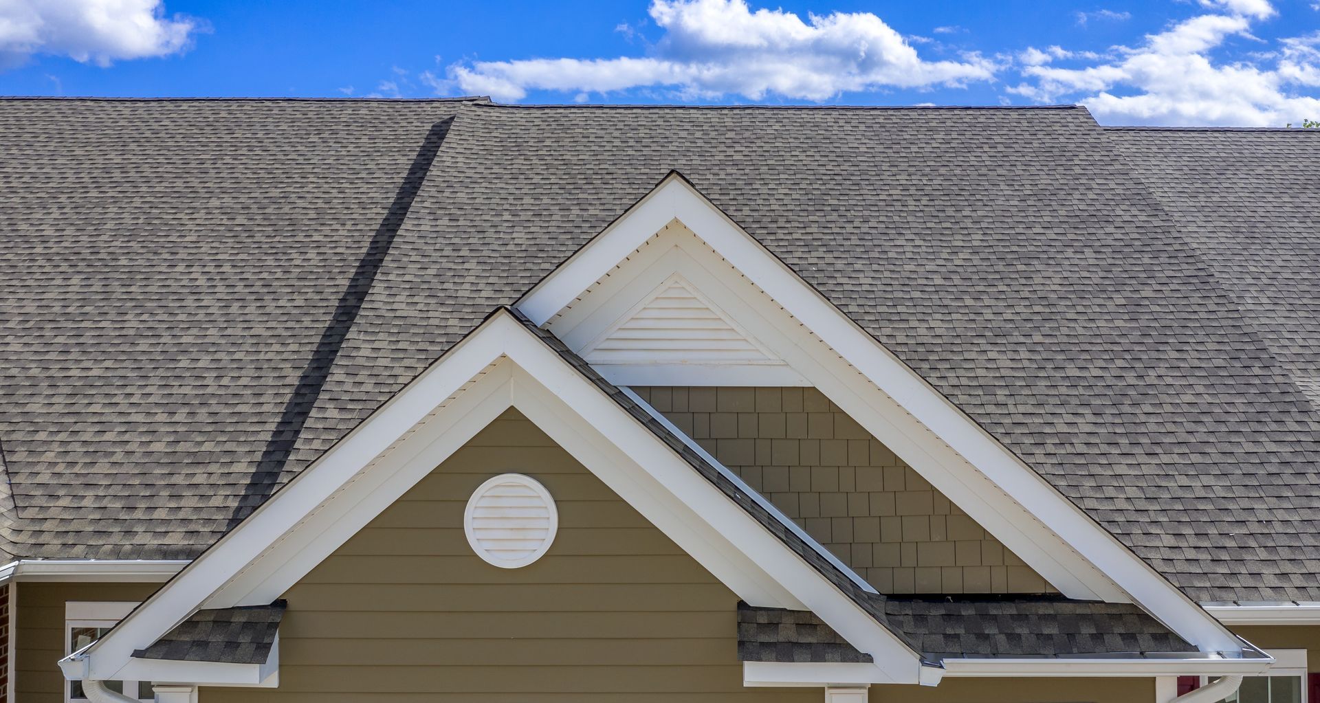 Vue détaillée d’une toiture de maison avec deux pignons, revêtement en tuiles grises, bardage beige et ciel bleu avec nuages.