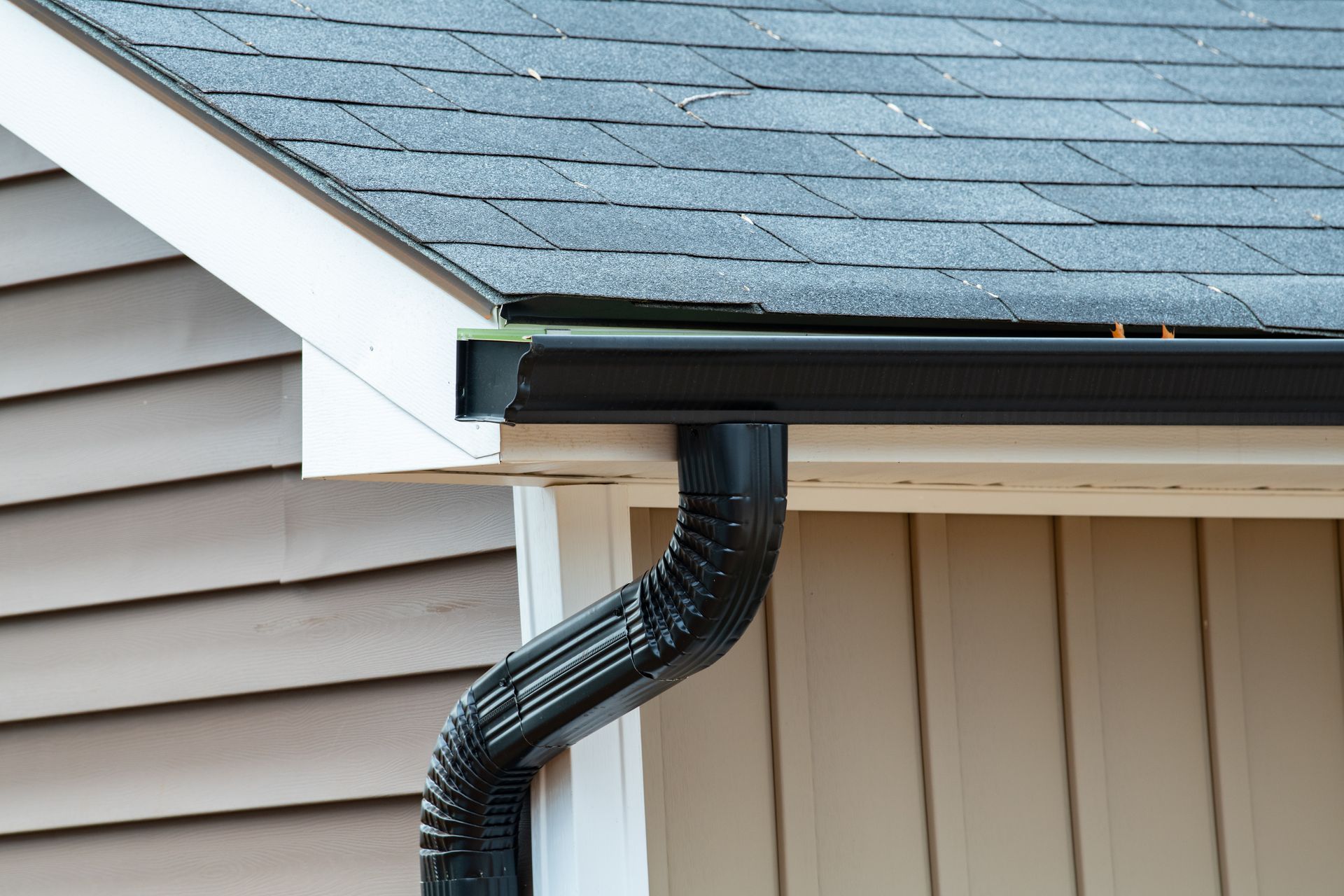 Descente de gouttière noire fixée sous l'avant-toit d'une maison, avec un revêtement extérieur beige et des bardeaux gris.