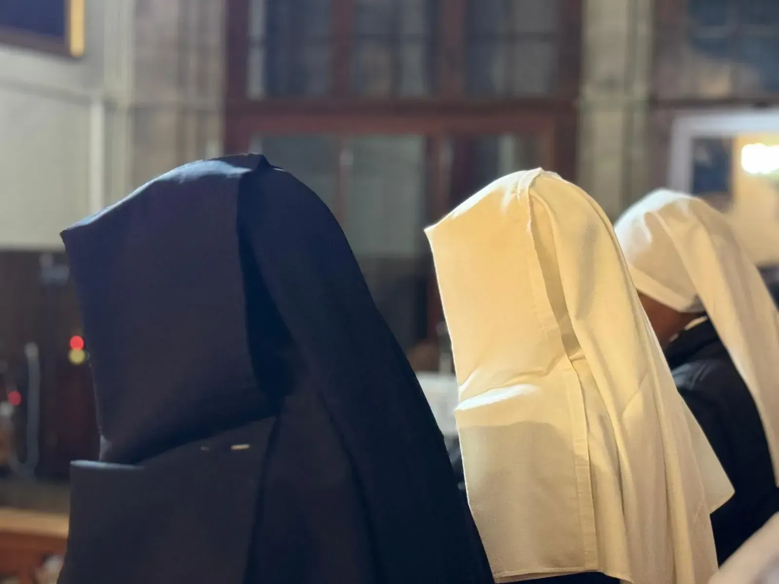 Tres monjas con hábitos, blancos y negros, dentro de una iglesia; capuchas que ocultan los rostros.