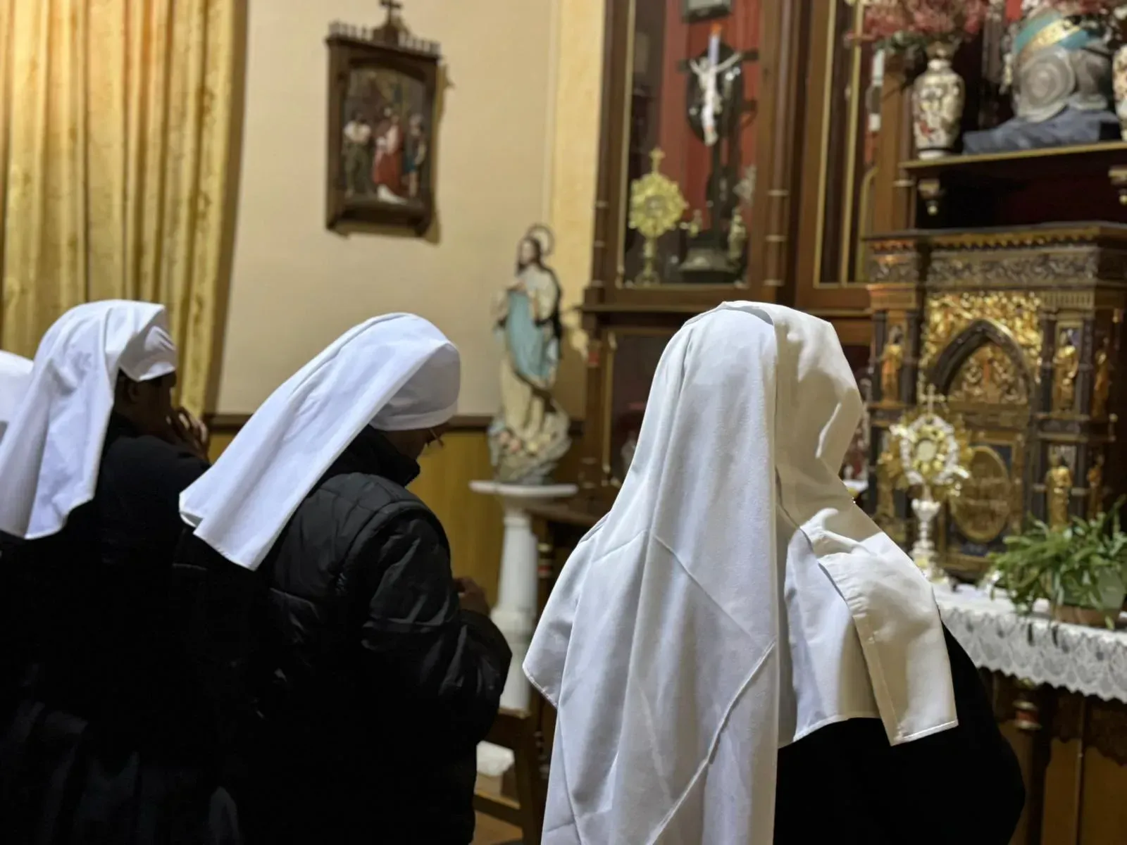 Monjas rezando en una capilla, de cara al altar. Llevan velos blancos y hábitos negros. La luz ilumina los objetos religiosos.