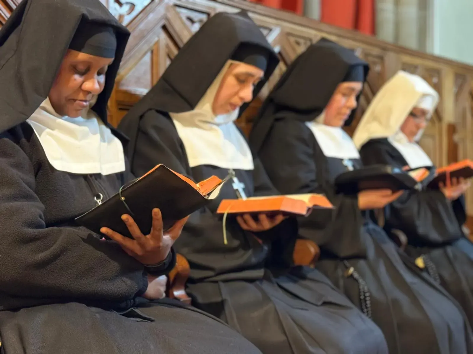 Monjas vestidas con hábitos negros leen libros sentadas en un banco de madera dentro de una iglesia.