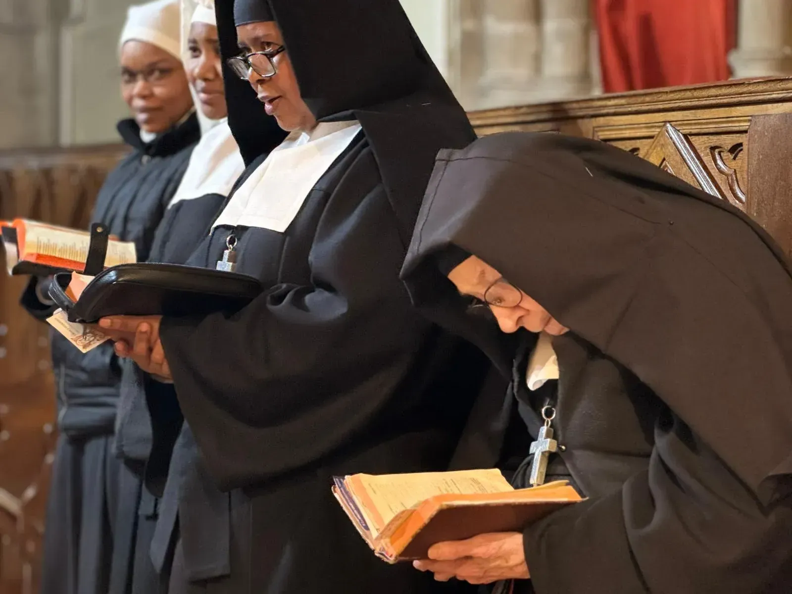 Monjas con hábitos negros leen libros en una iglesia. Se ve un banco de madera.