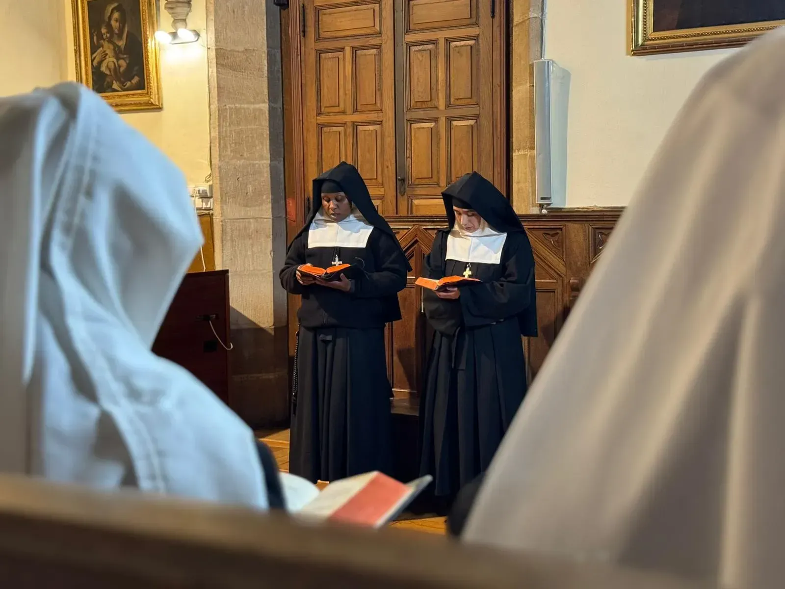 Dos monjas vestidas con hábitos negros sostienen velas encendidas en una iglesia con paneles de madera.