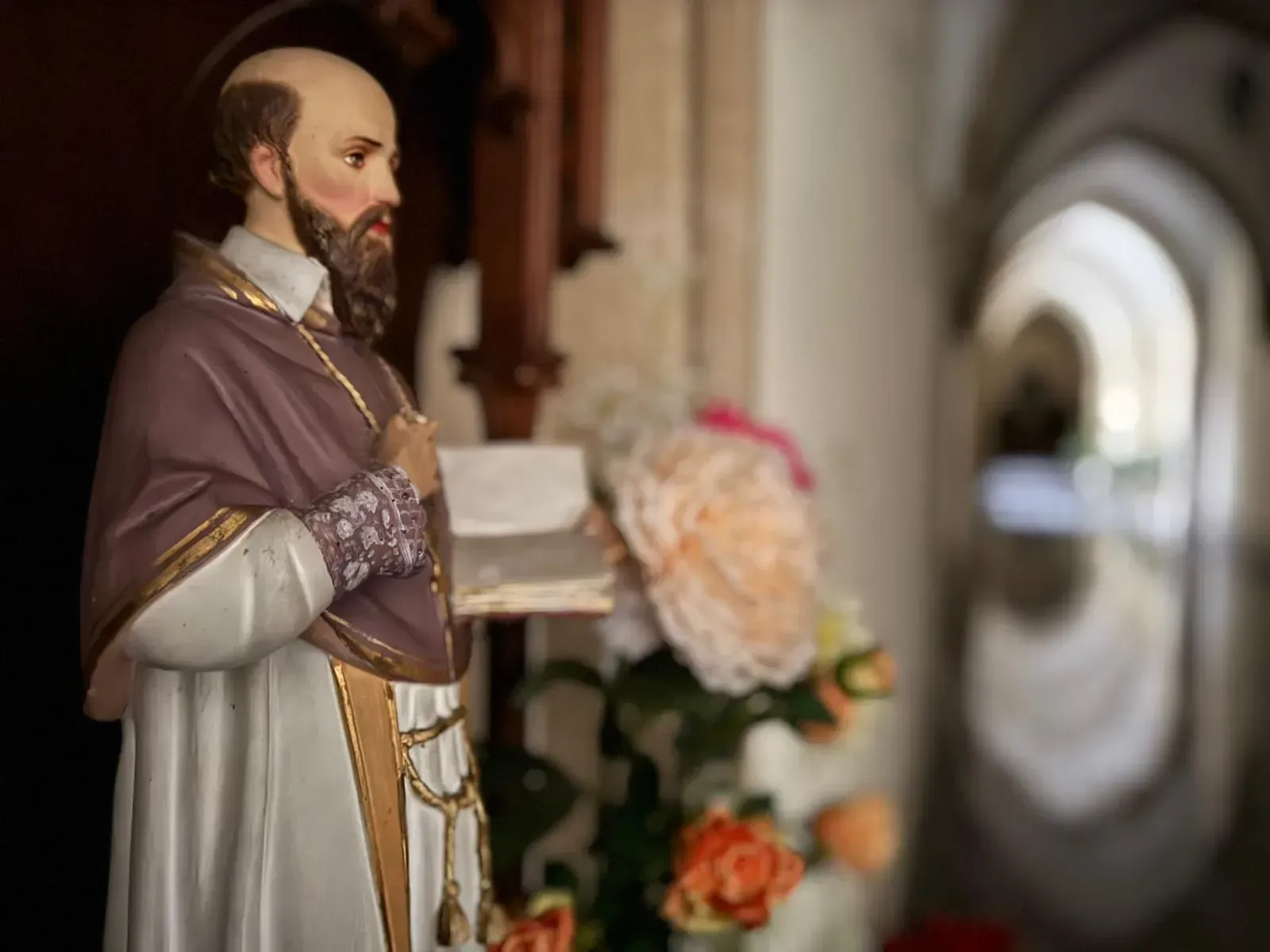 Estatua de un hombre con vestimenta religiosa, sosteniendo un libro, con un arco de fondo borroso.