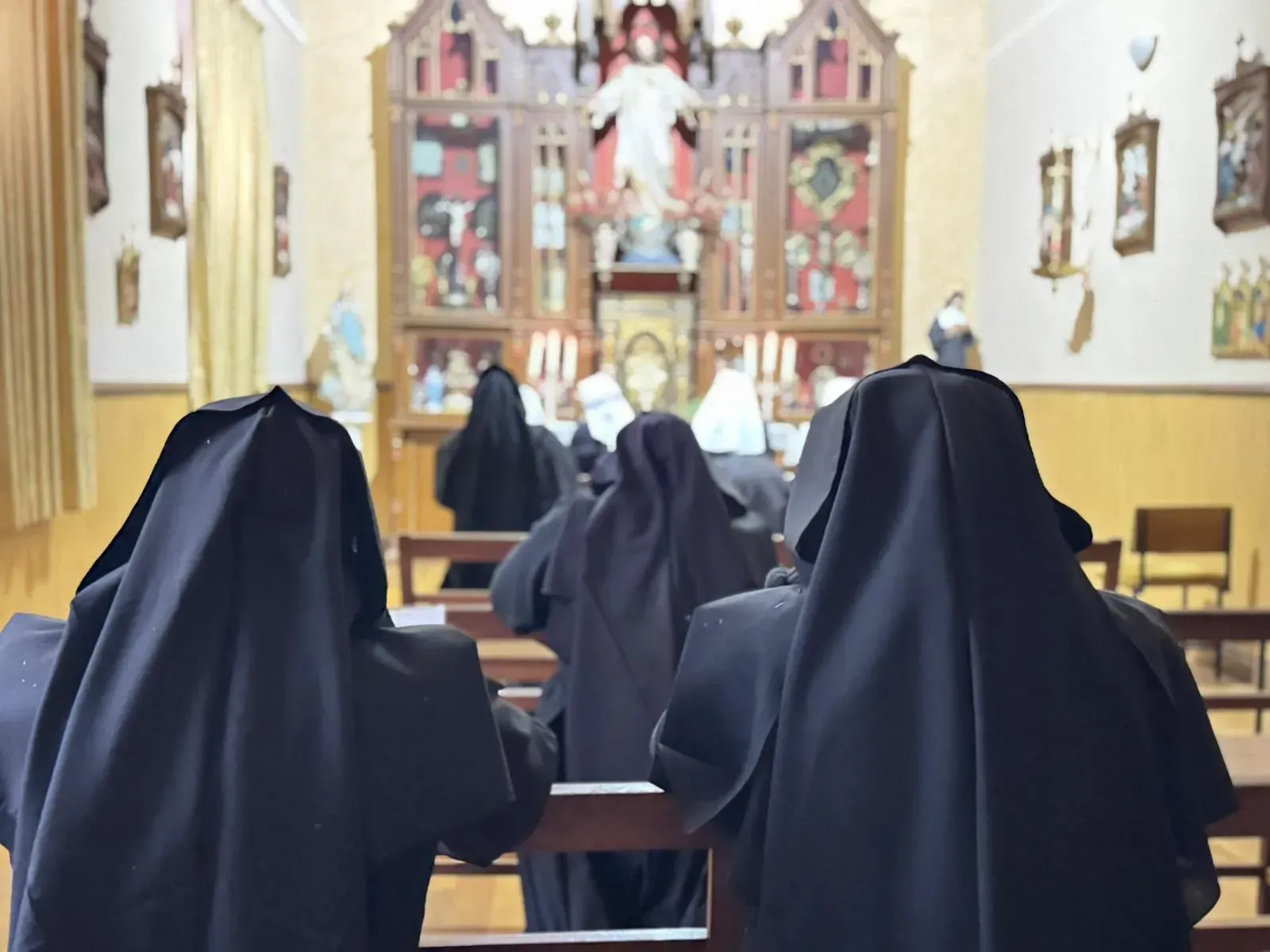 Monjas con hábitos negros rezando en una capilla, frente a un altar con obras de arte religioso.